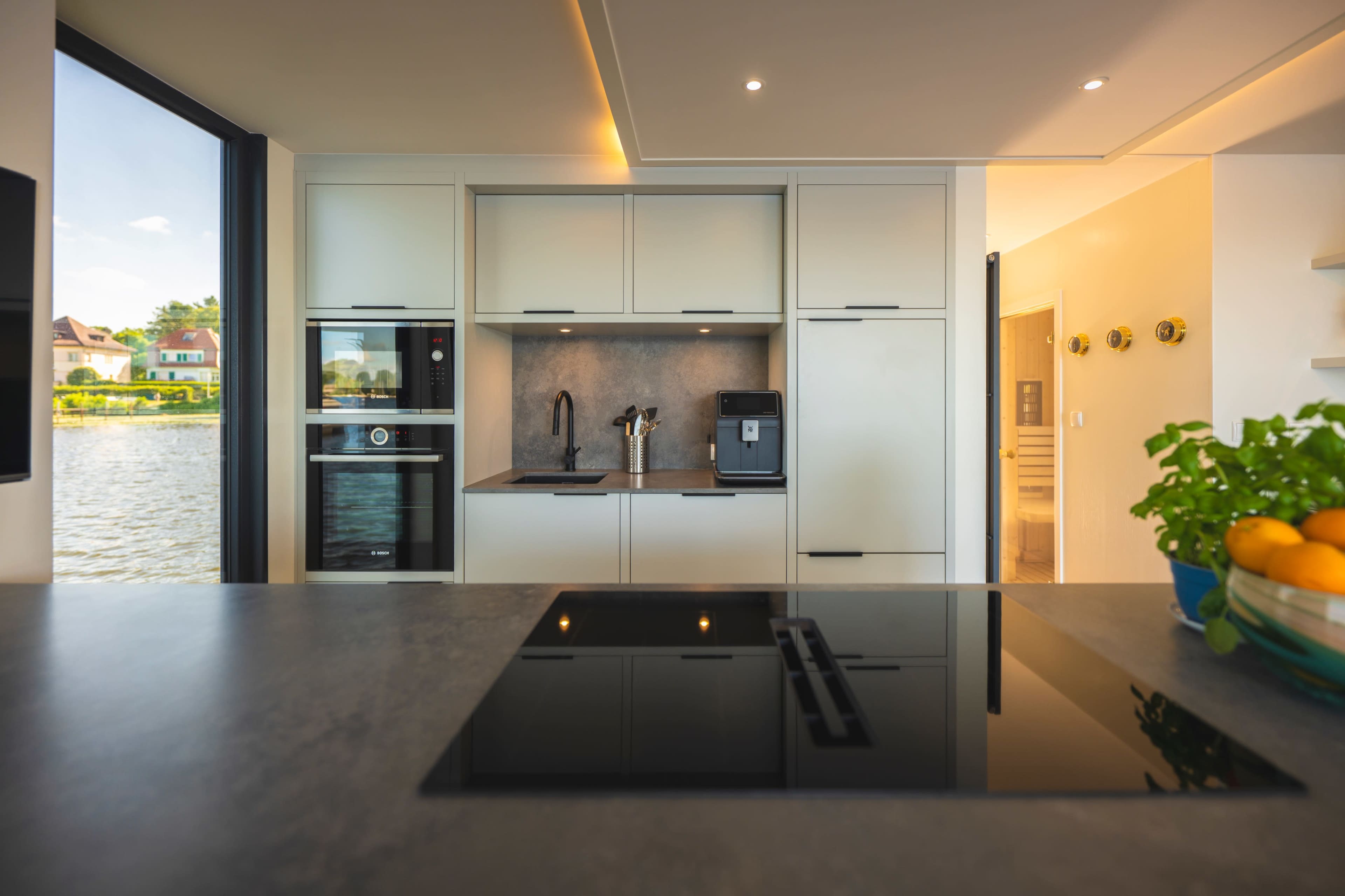 A modern kitchen features a sleek design with built-in appliances, including an oven, coffee maker, and an induction cooktop, overlooking a view of water and greenery outside.