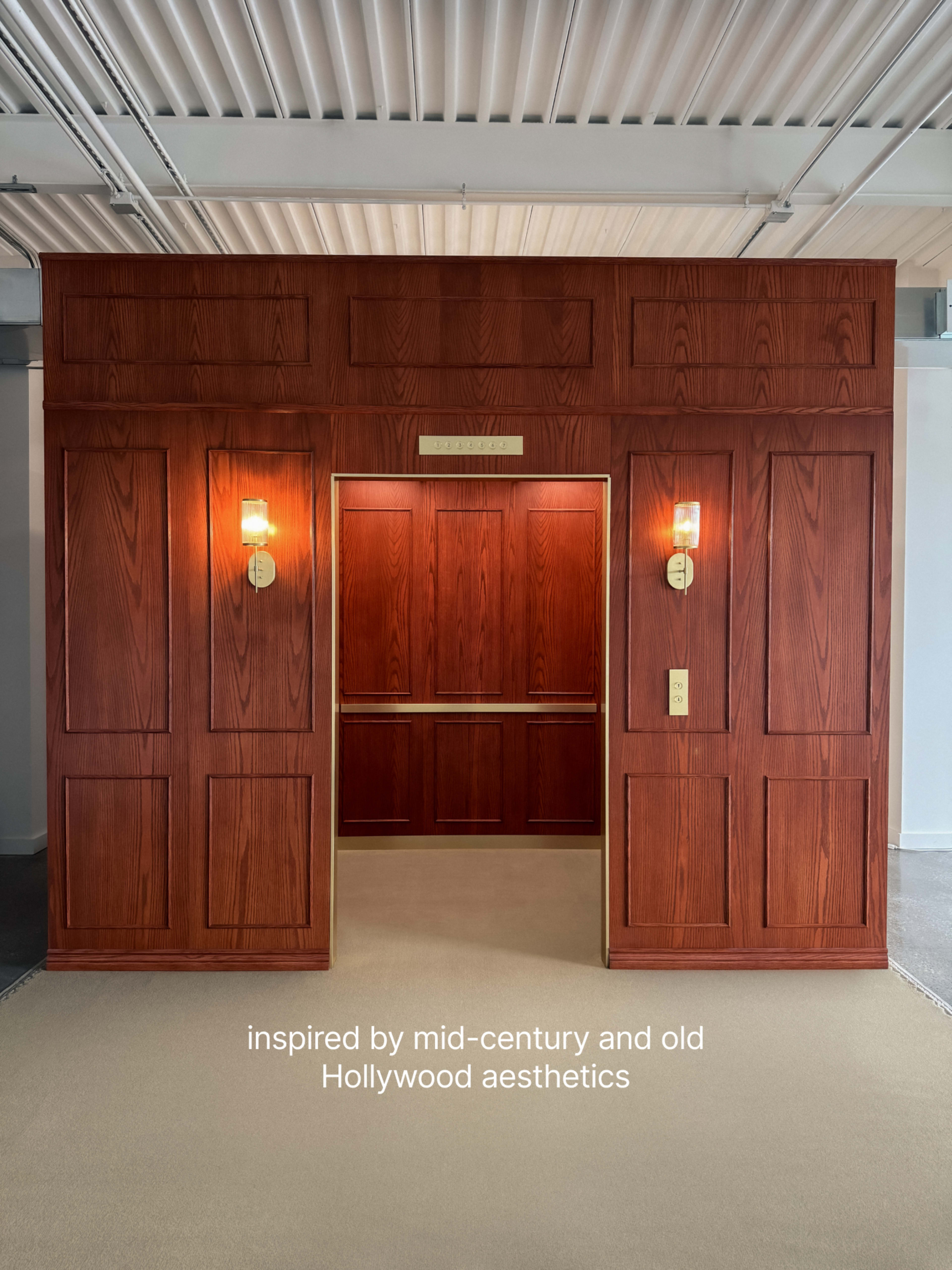 The image shows an elevator entrance designed with a mid-century and old Hollywood aesthetic, featuring wooden paneling and vintage-style lighting fixtures.