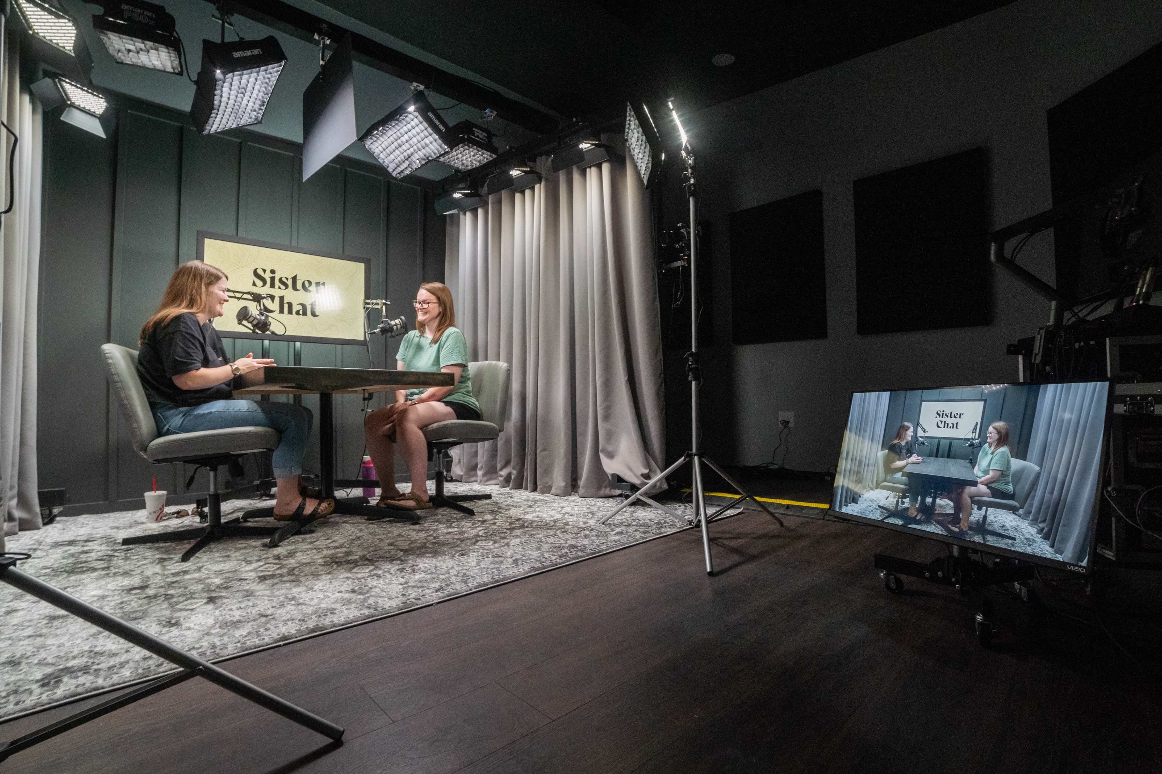Two women are seated at a table in a well-lit studio, engaging in conversation while a camera captures the scene.