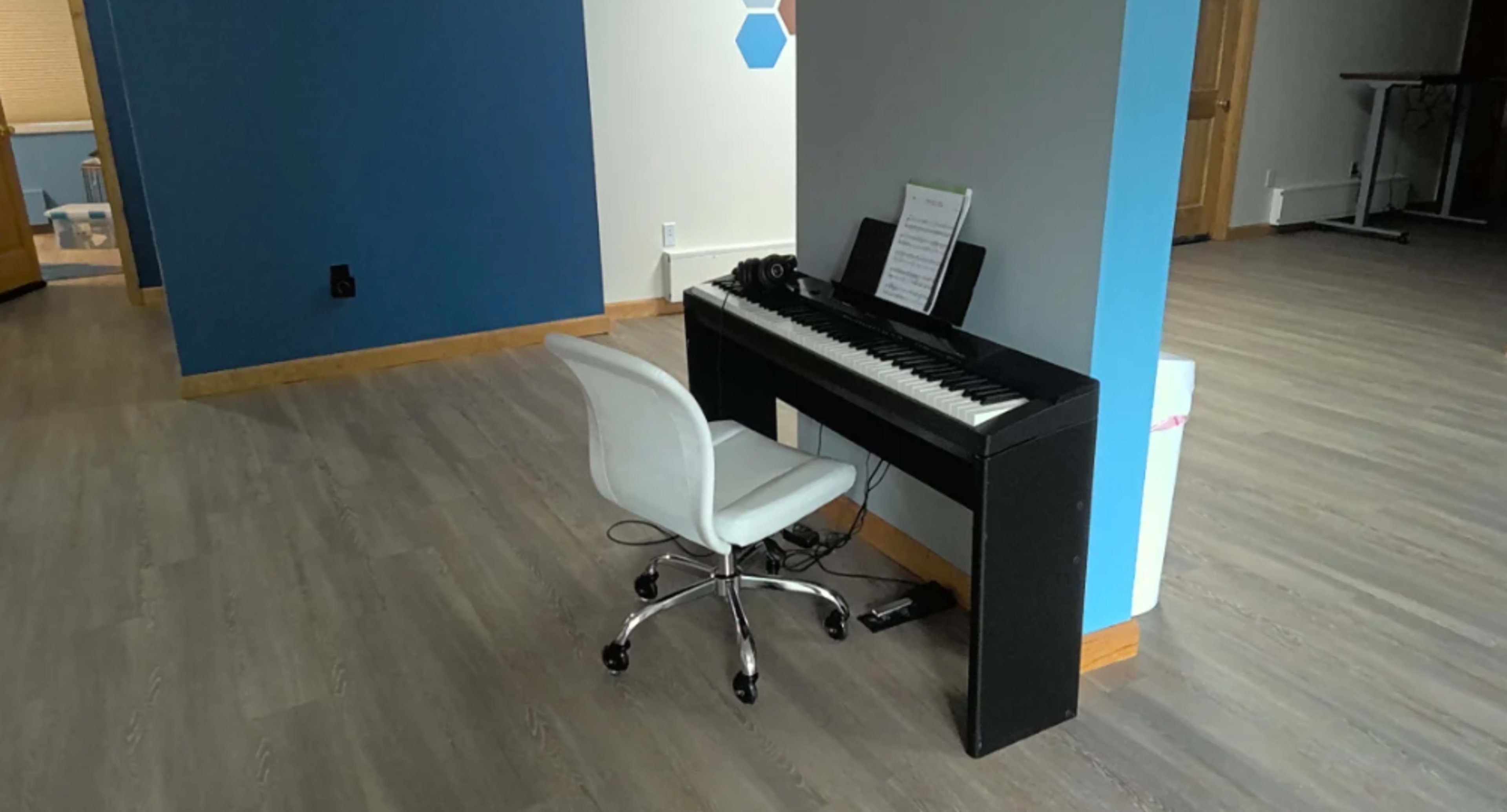A black piano with sheet music and a white chair is positioned against a blue and gray wall in a spacious room with wooden flooring.