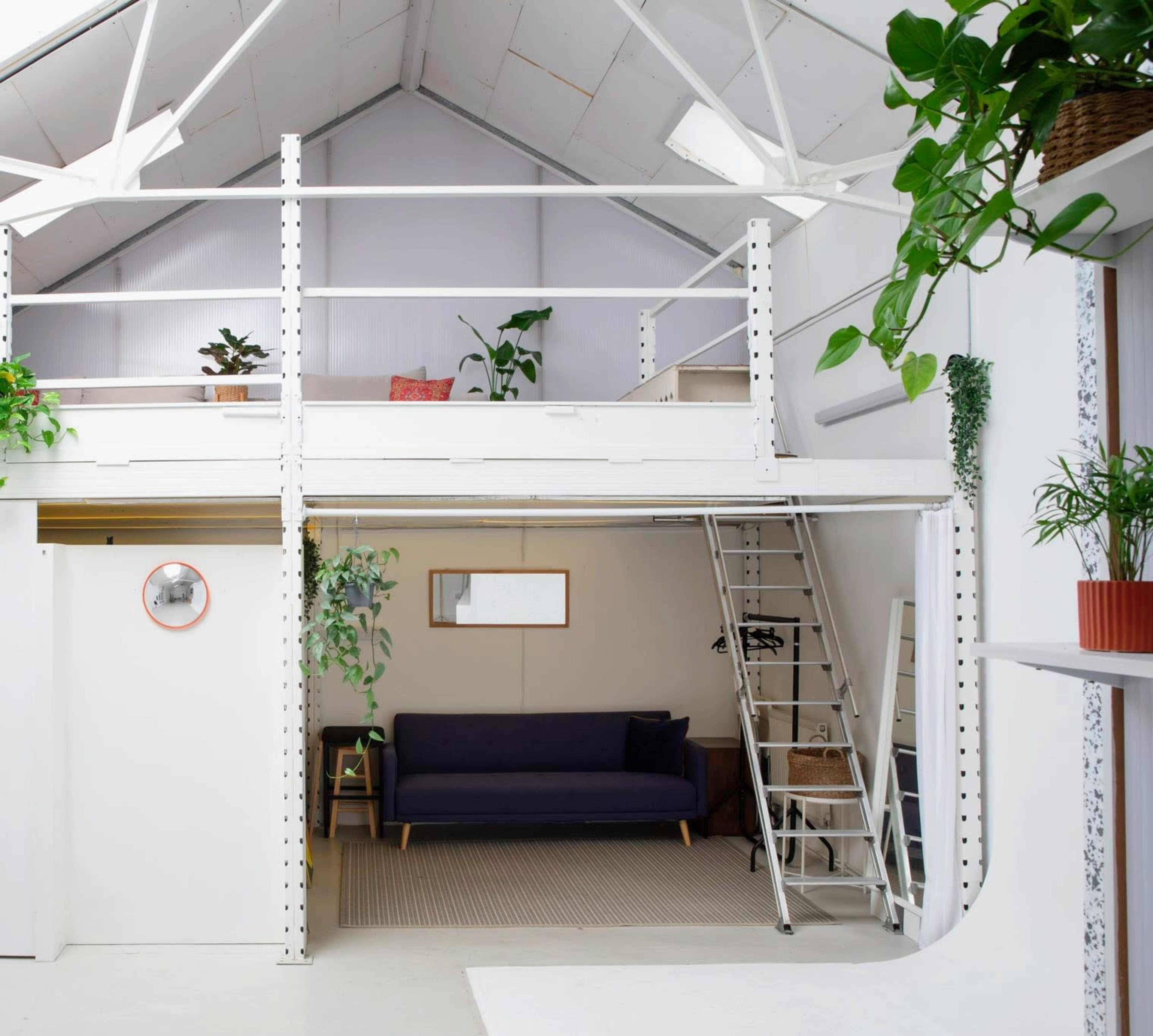 The image shows a modern loft space with a mezzanine, featuring a sofa on the lower level and plants on both levels.