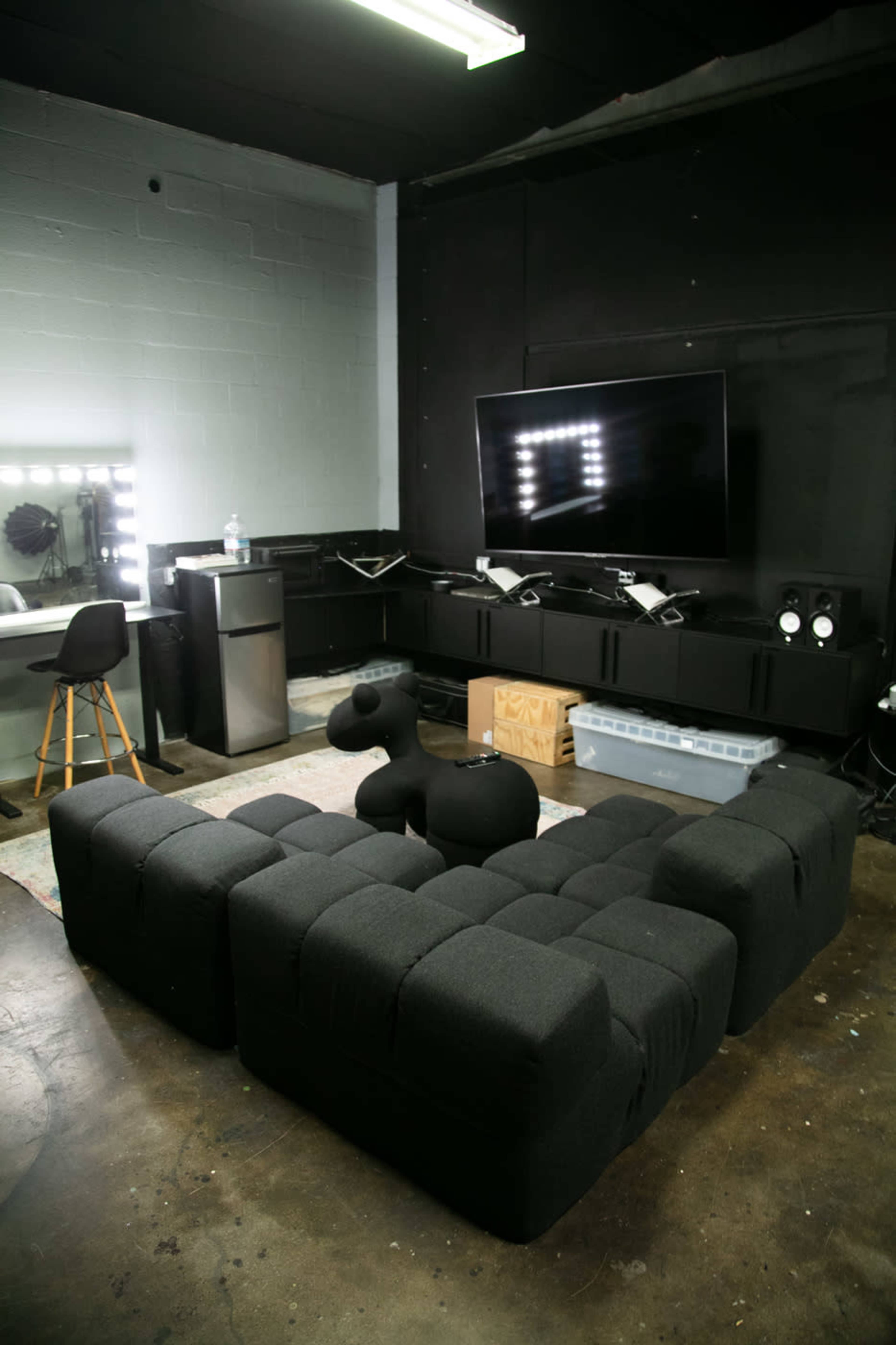 A modern lounge area with a black fabric modular sofa, a large television on the wall, a small kitchenette with a refrigerator, and a stylish black stool.