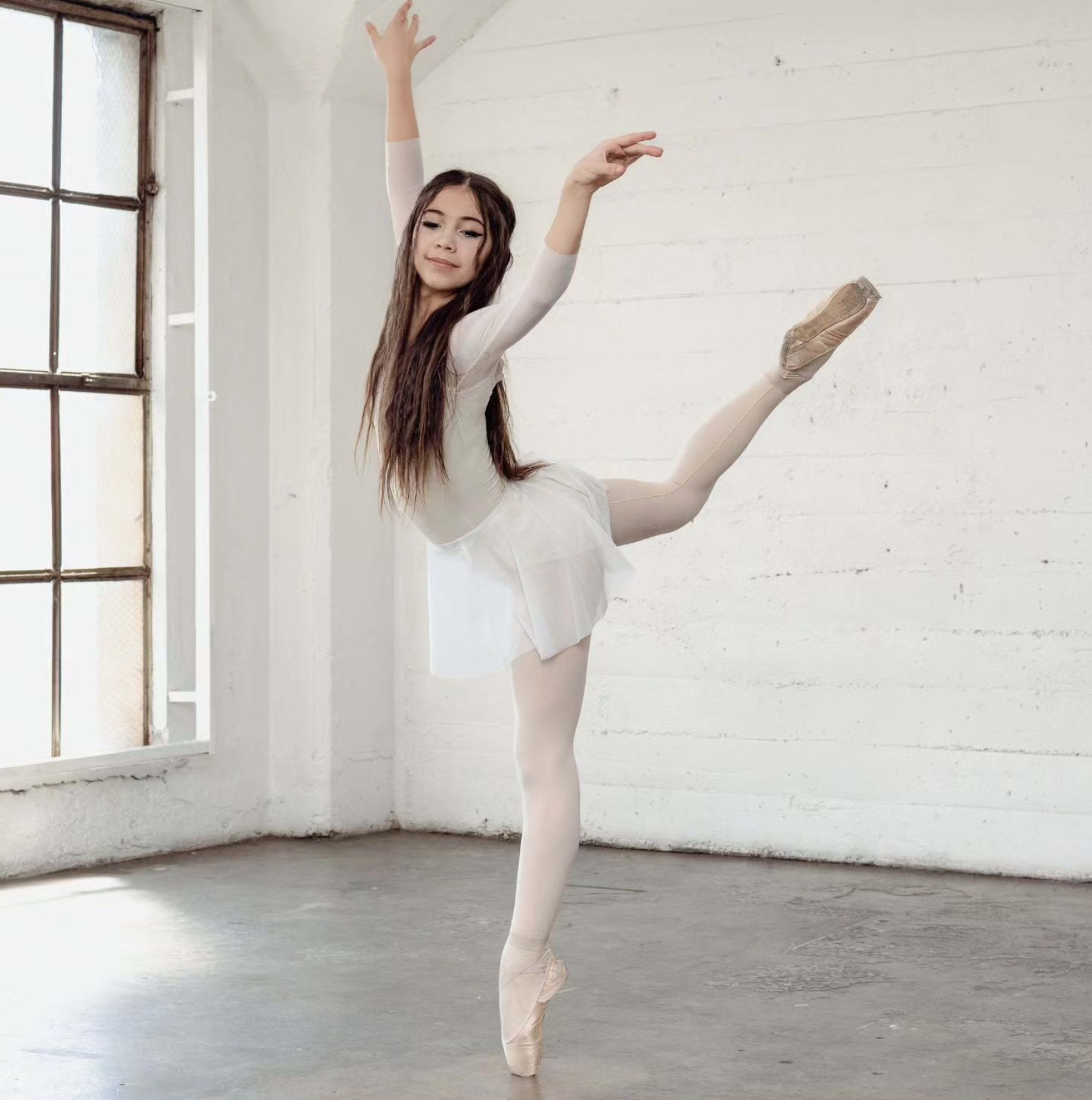 A young ballerina is posed en pointe in a bright, minimalist studio with large windows.