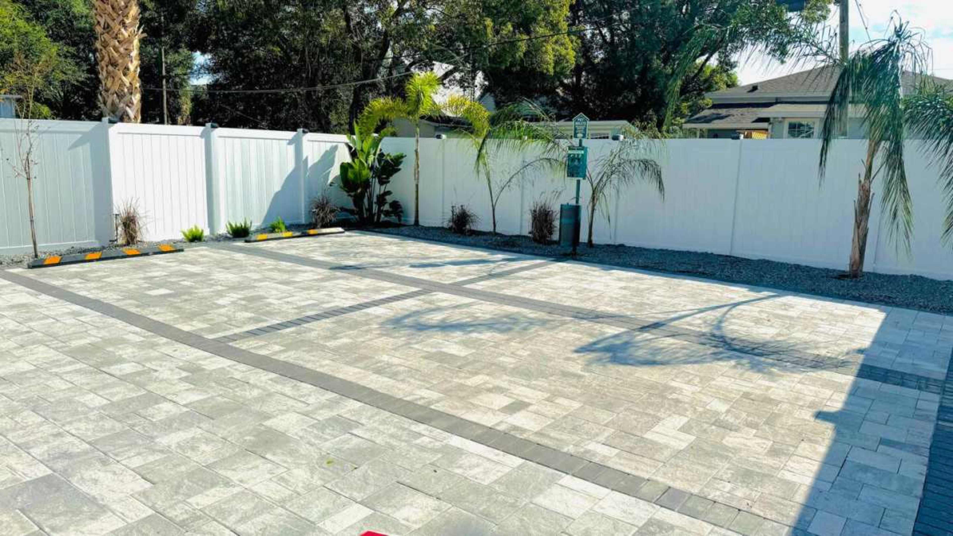 The image shows a landscaped outdoor area with a paved surface, surrounded by fences and various plants.