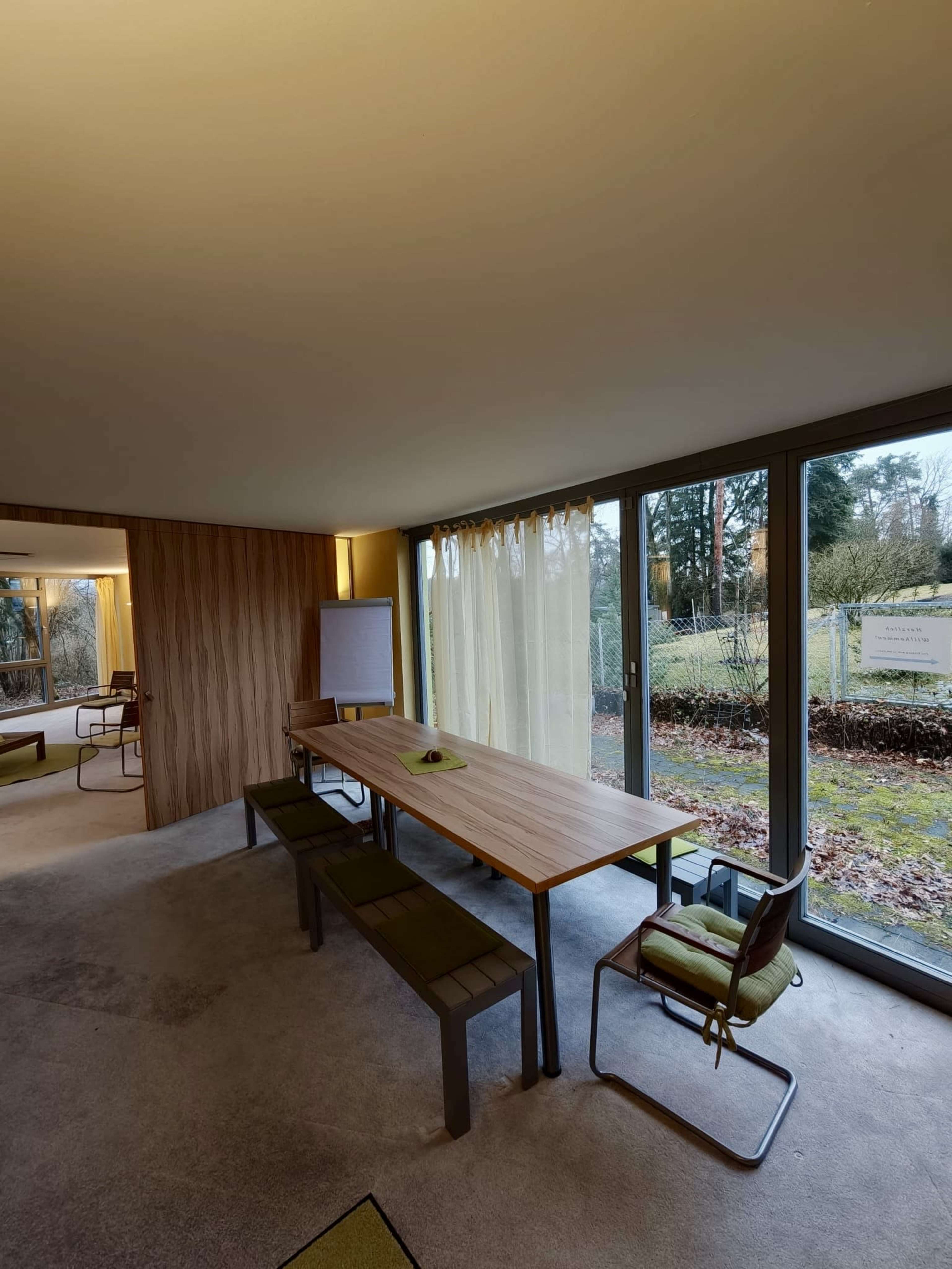 The image shows a modern meeting room featuring a long wooden table with chairs, large windows overlooking greenery, and a whiteboard on one side.