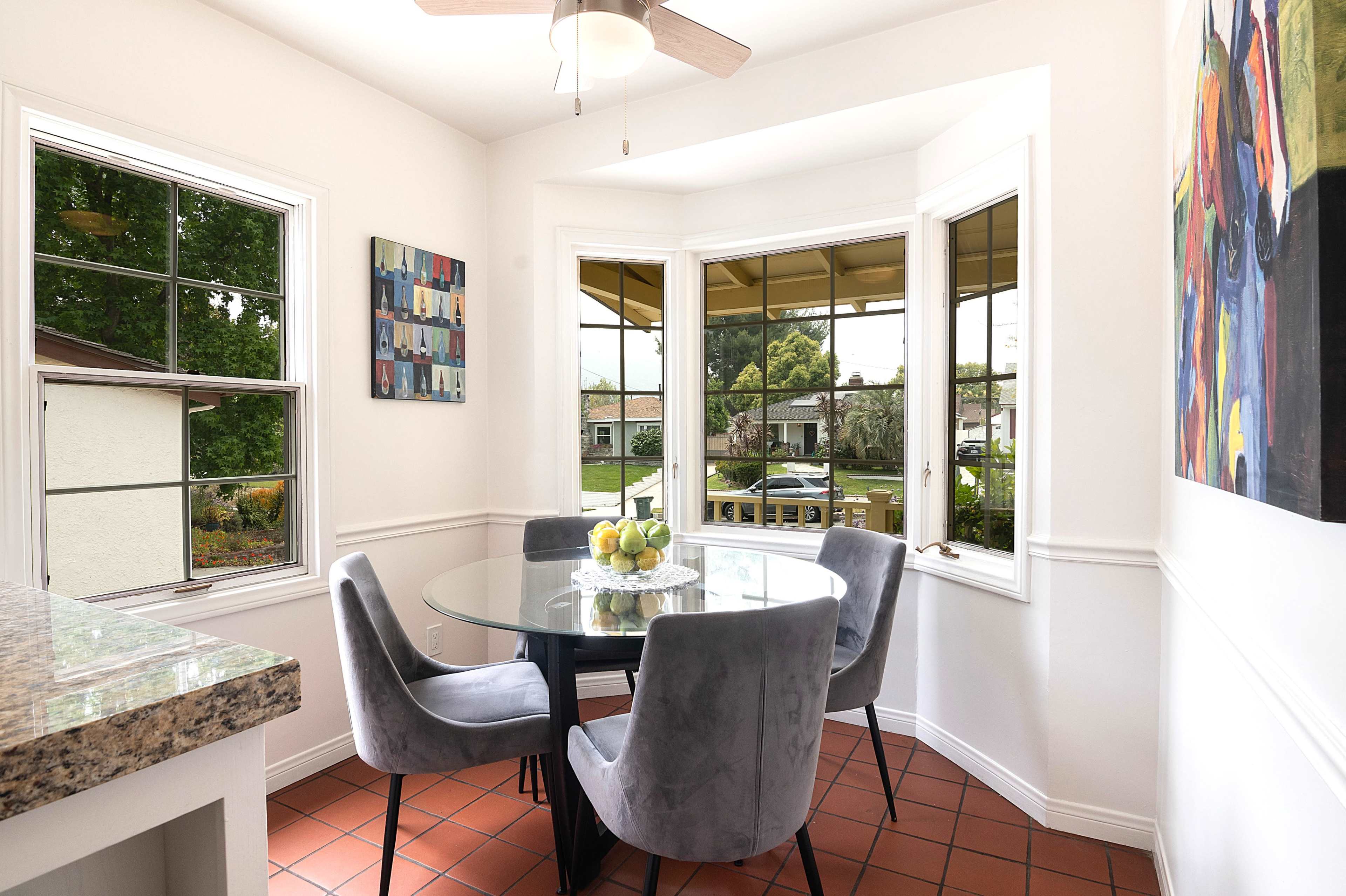 A round dining table with gray chairs is positioned near large windows in a bright kitchen area, showcasing a view of the outdoors.