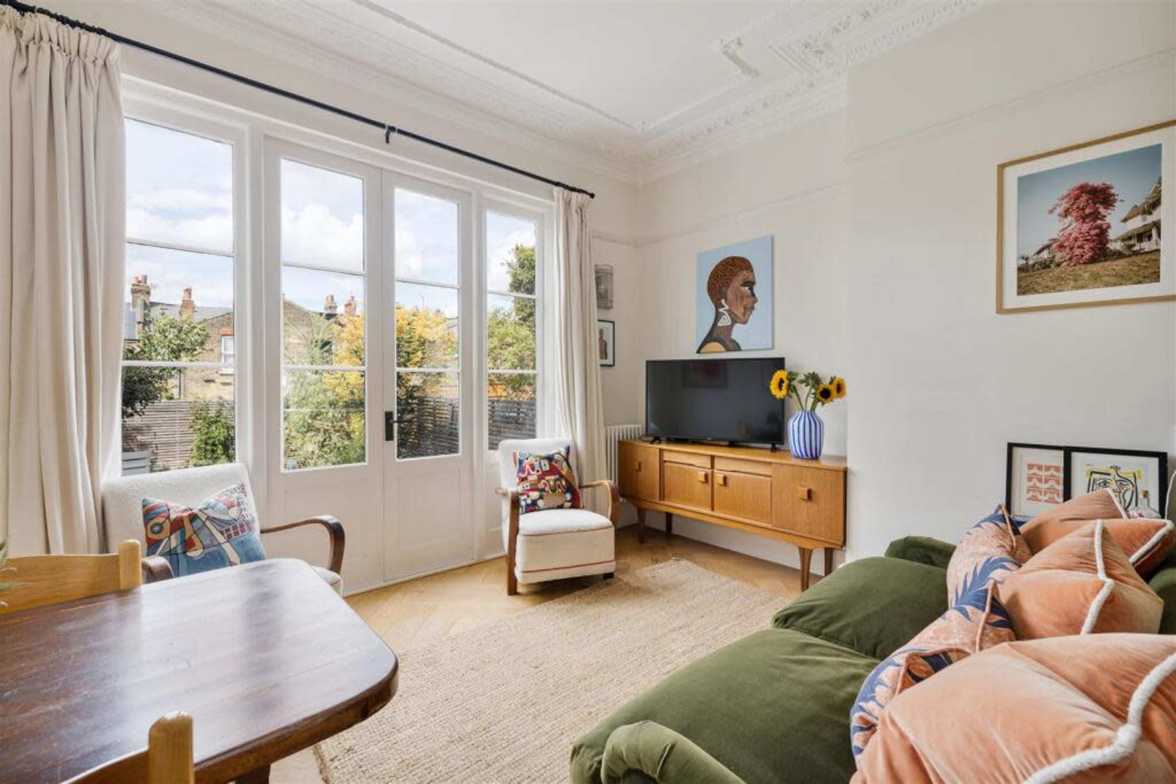 The image shows a cozy living room with large windows, a television on a wooden cabinet, a green sofa, and a small dining table.