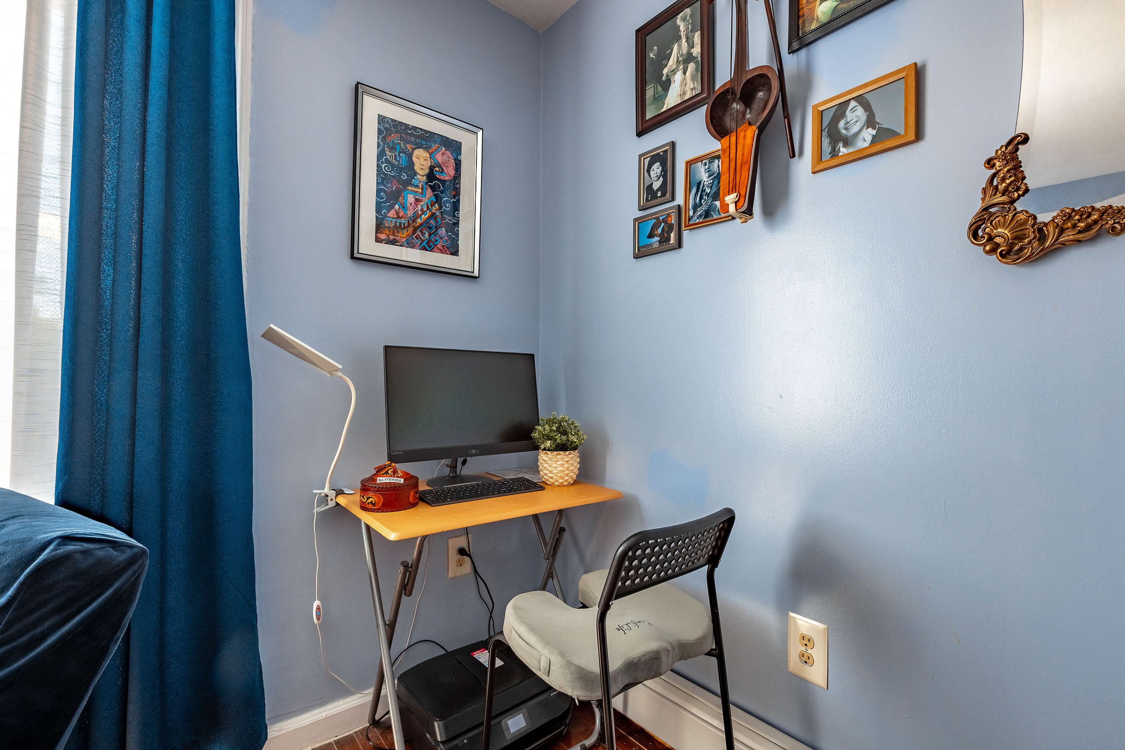 A small desk with a computer and chair is situated in a corner of a blue-walled room adorned with framed artwork.