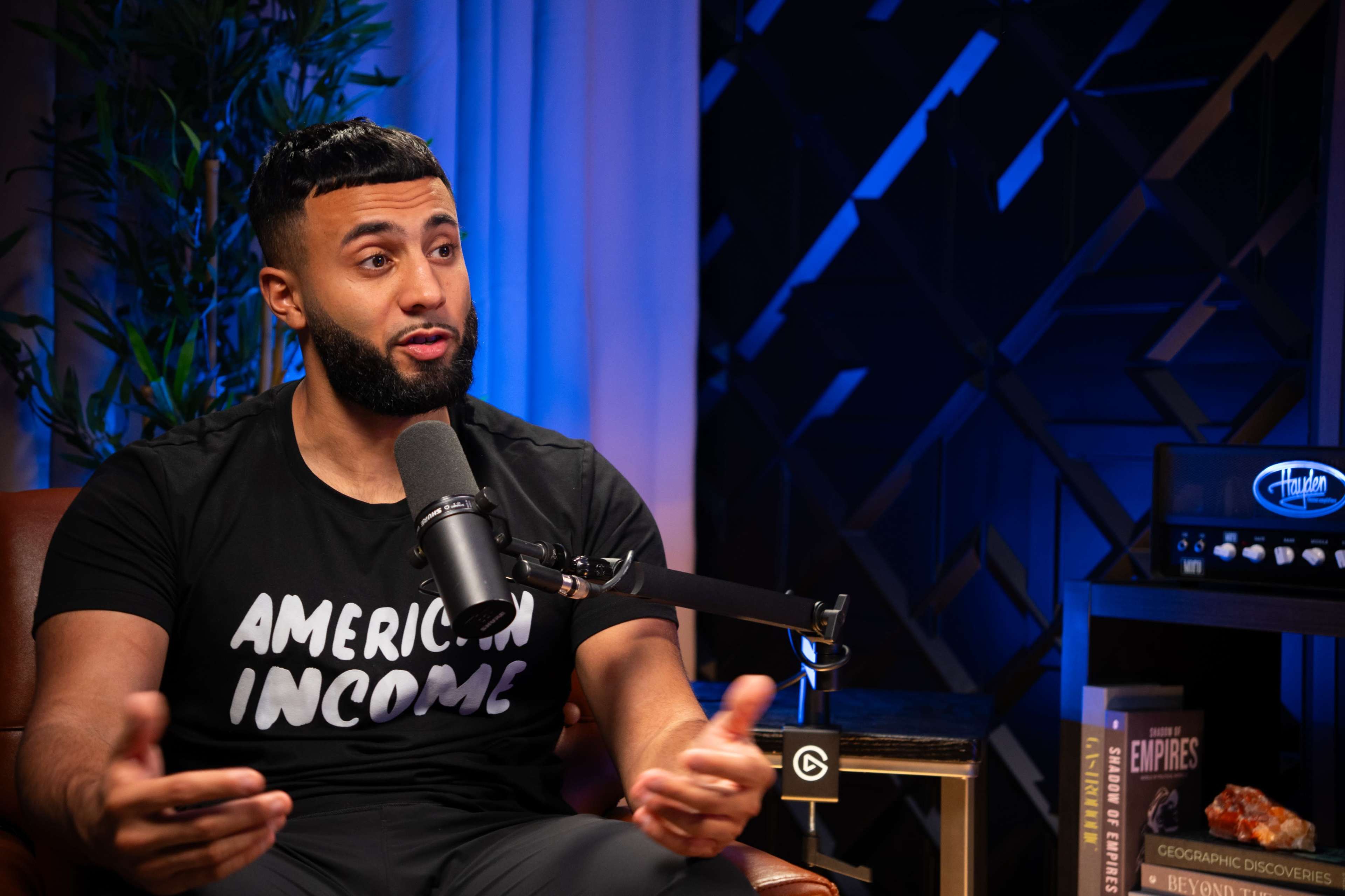 A man in a black T-shirt is speaking into a microphone while sitting in a well-lit studio equipped with modern decor.