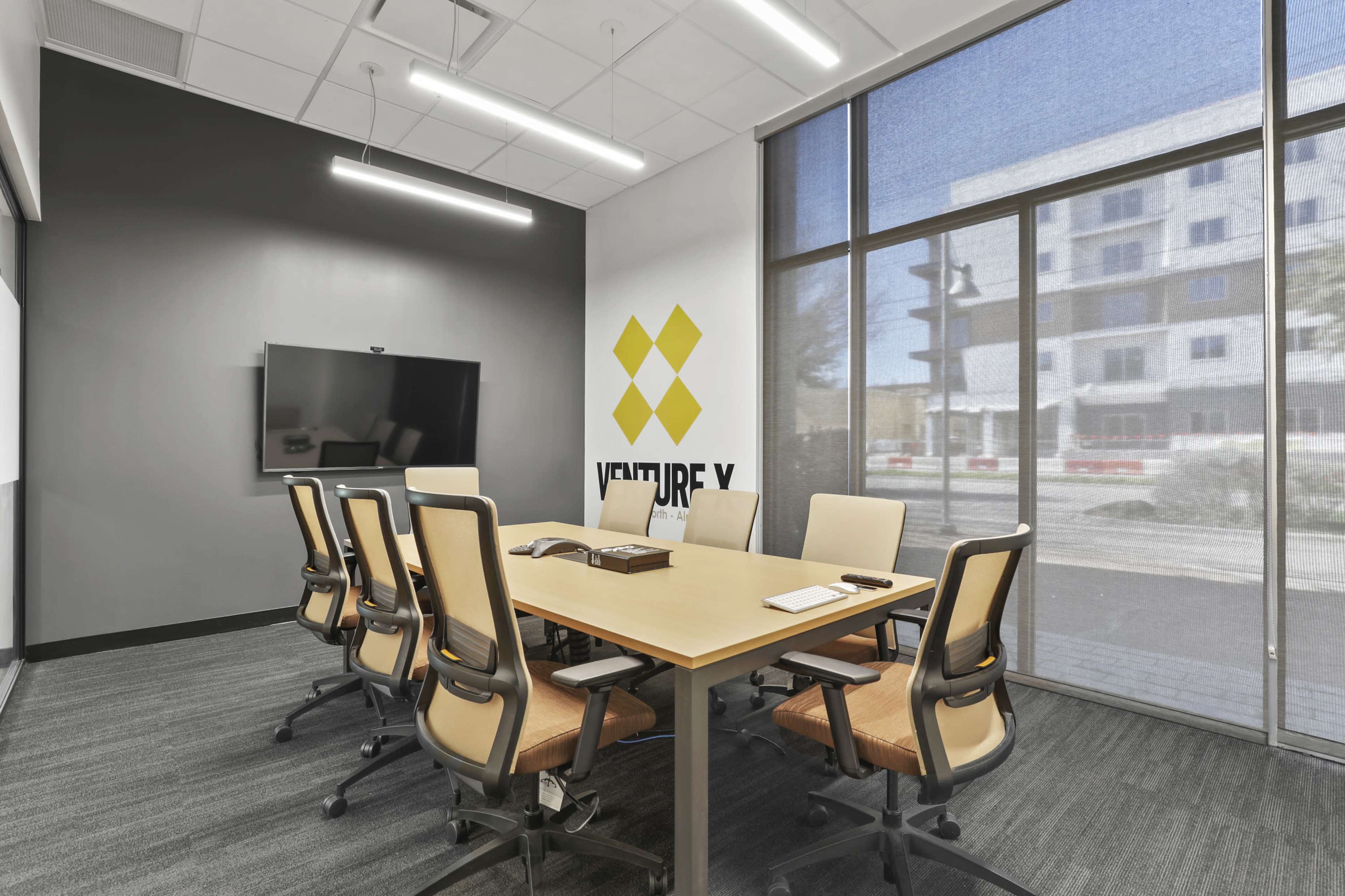 A modern conference room features a large table surrounded by eight ergonomic chairs, a black screen mounted on the wall, and a logo displayed on the gray backdrop.
