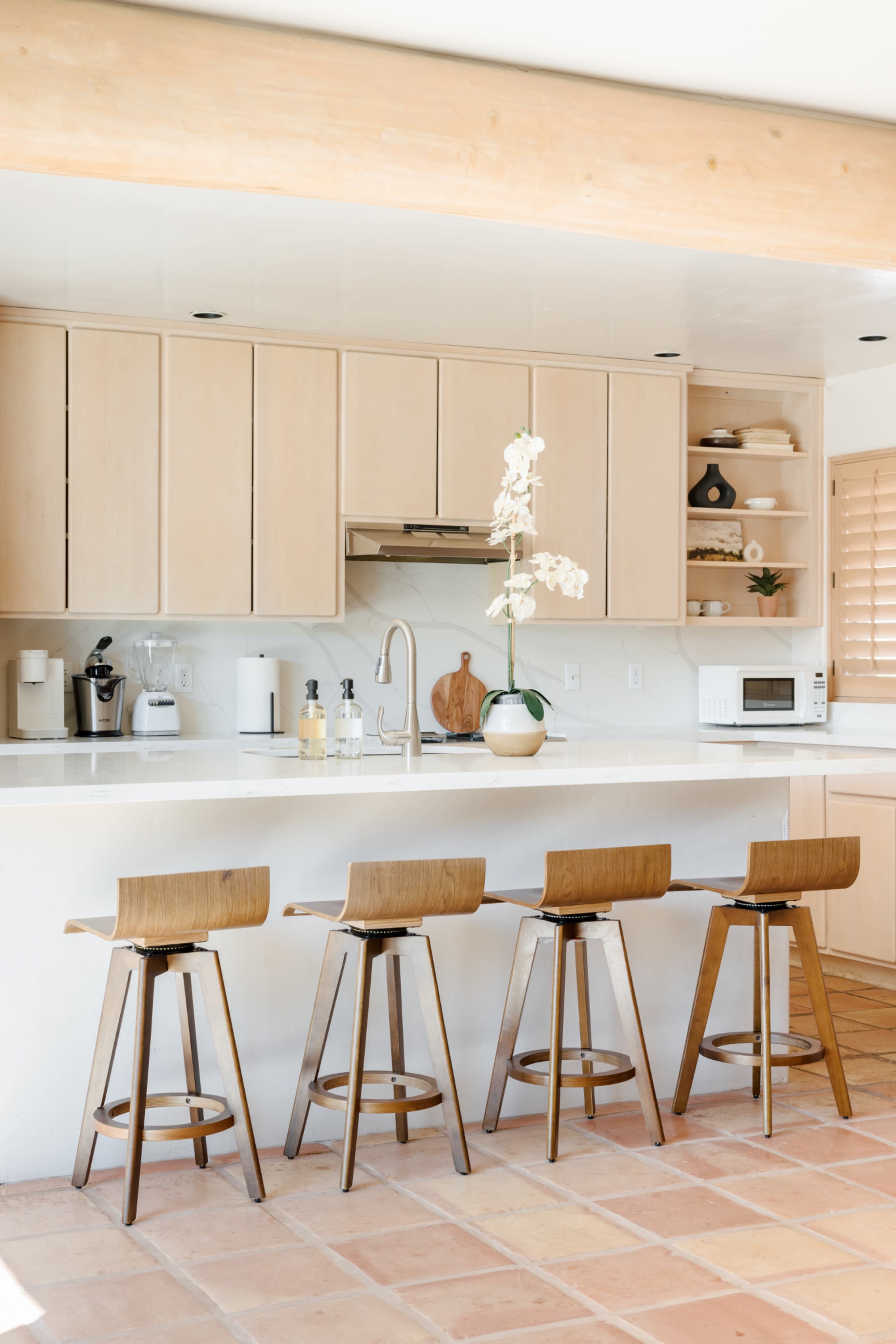 A minimalist kitchen features light wooden cabinetry, a countertop with a sink, and four wooden bar stools arranged in front.