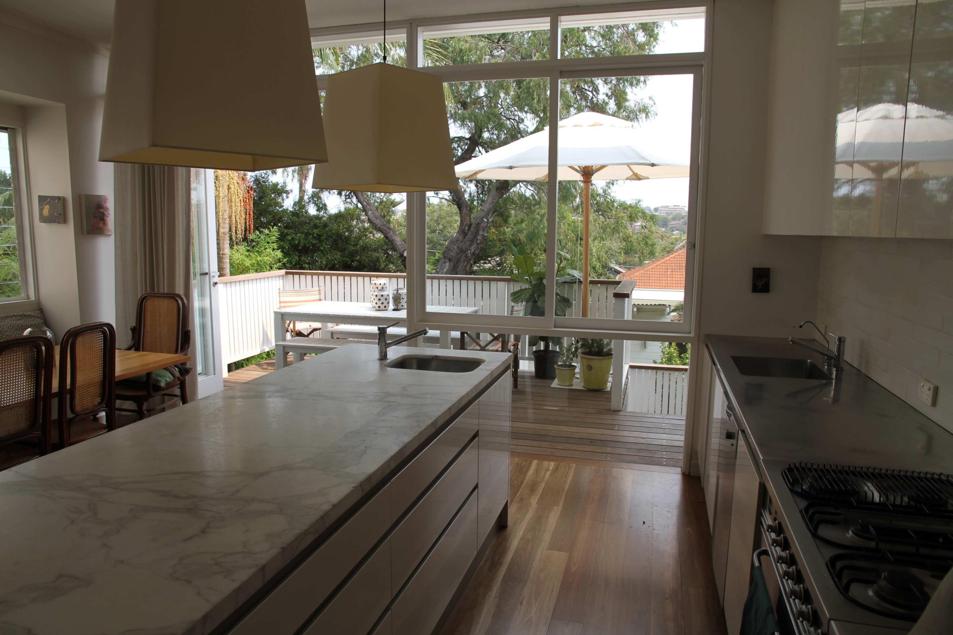 A modern kitchen featuring a large marble island and open windows that overlook a deck with outdoor seating and an umbrella.