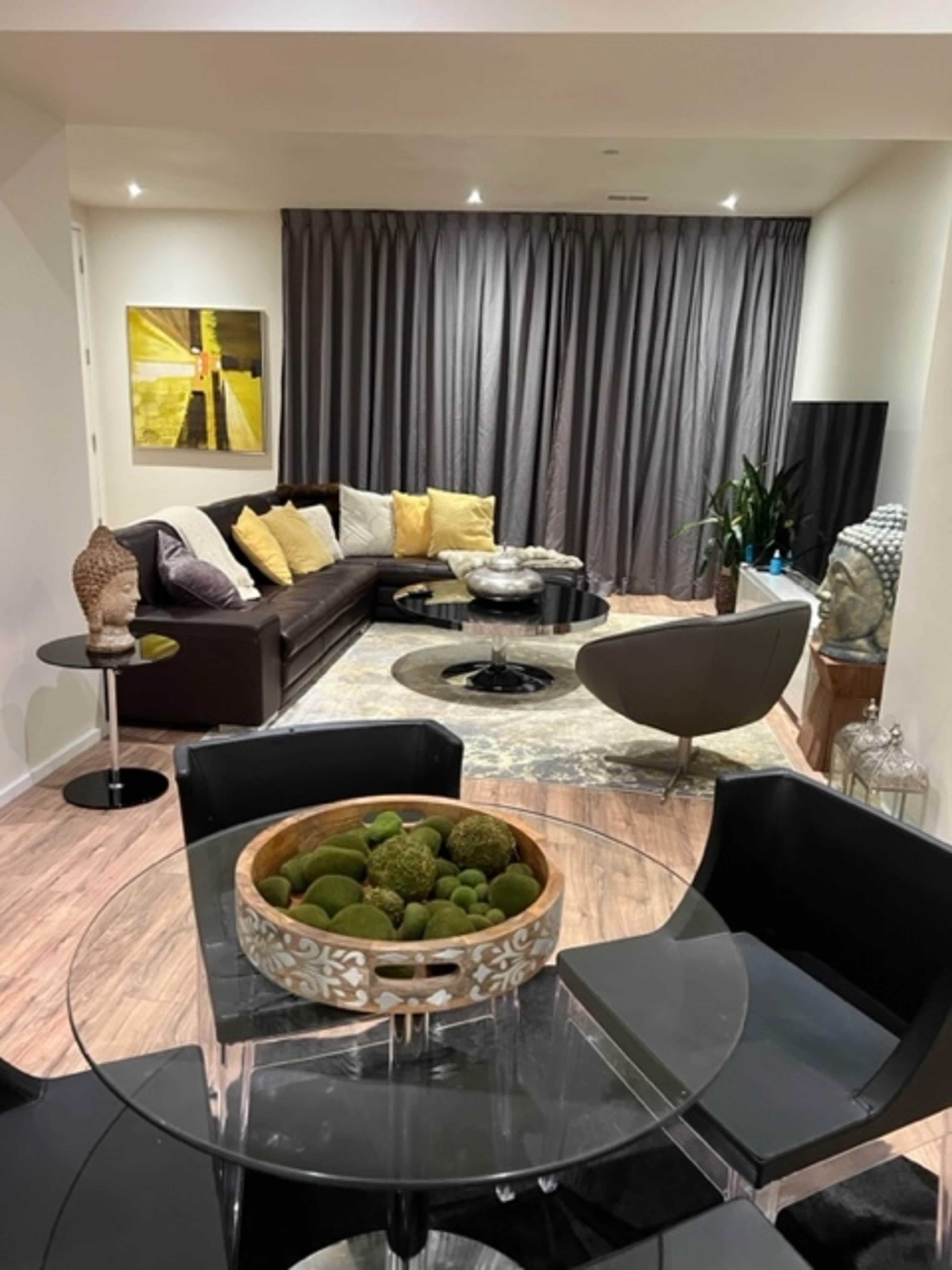 A modern living area with a sectional sofa, a circular chair, a glass dining table, and decorative elements such as a bowl of green ornaments and curtains.