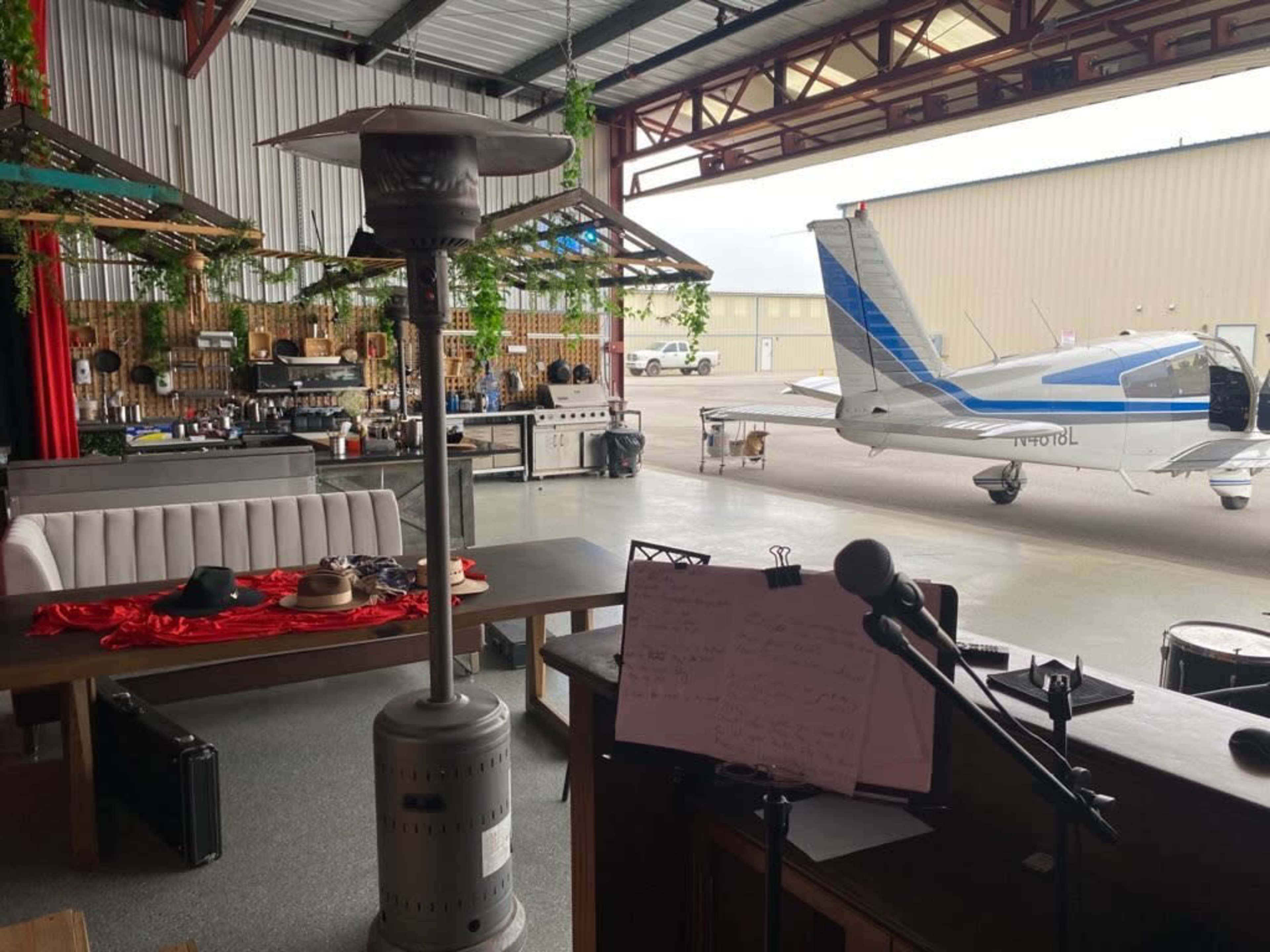 An open-air space featuring a bar area with seating, stage equipment, and a small aircraft parked outside a hangar.