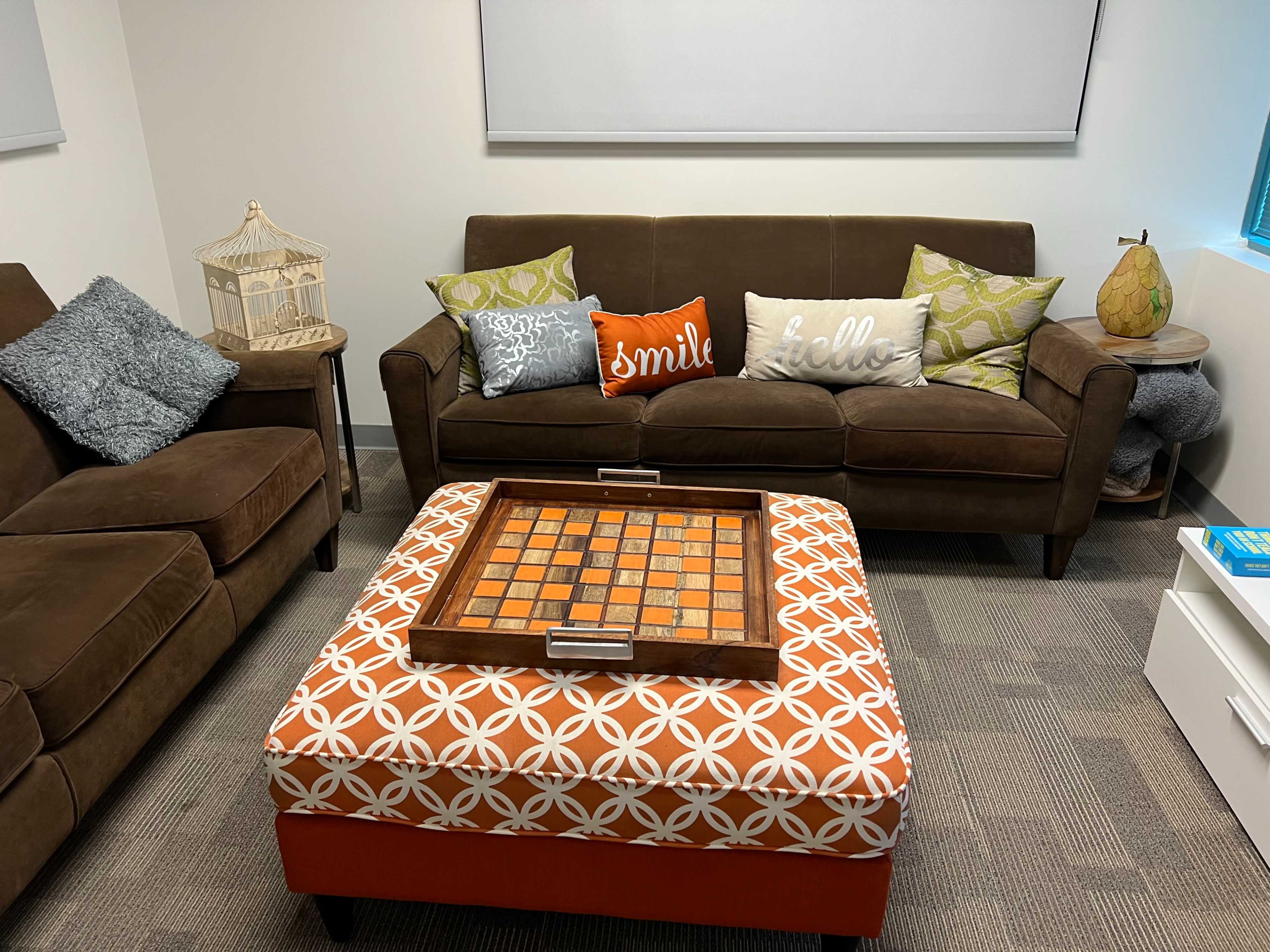 A cozy seating area with a brown sofa and armchair, adorned with decorative pillows, and a patterned ottoman positioned at the center with a wooden game board on top.