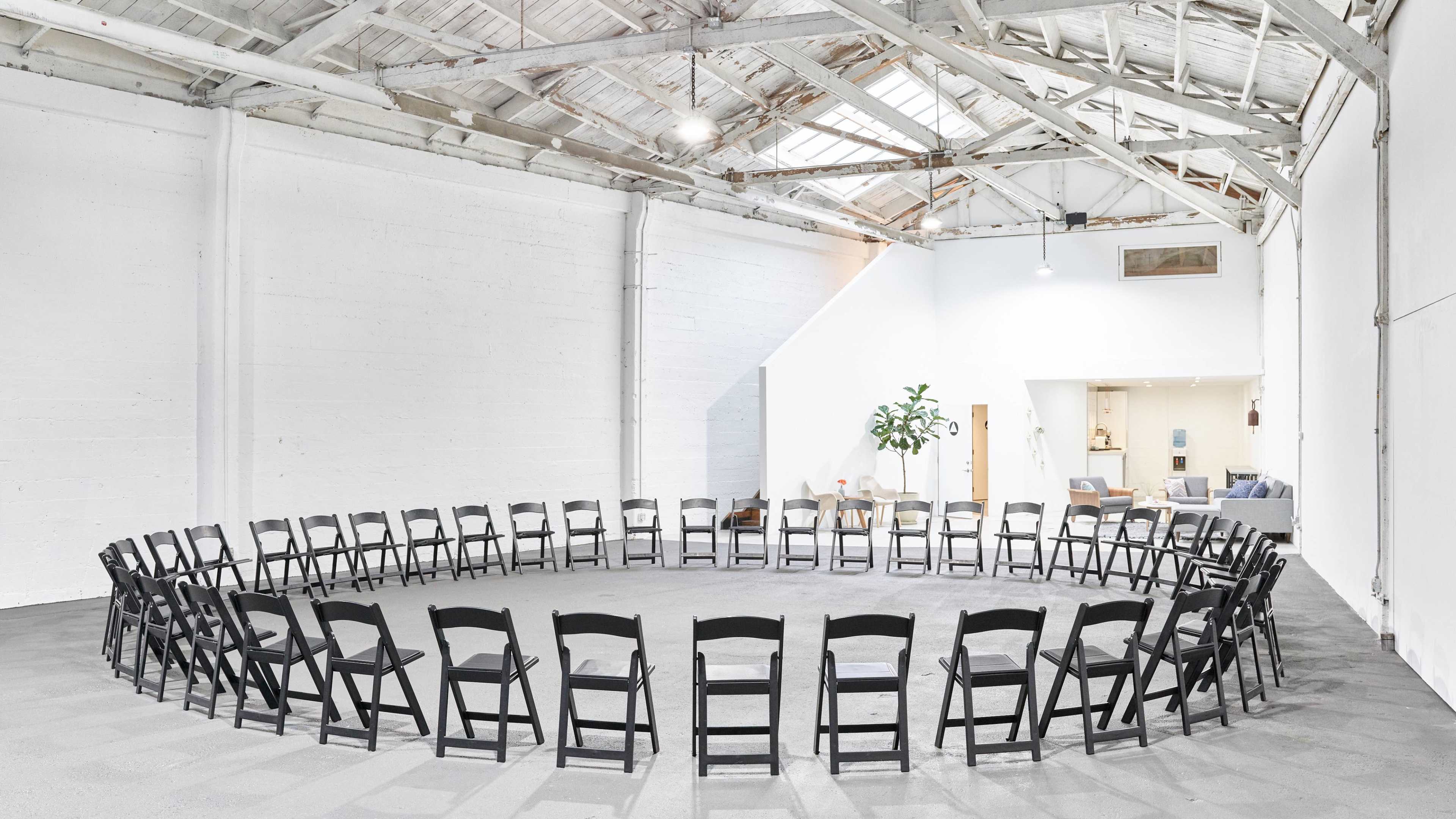 A circular arrangement of black chairs is set up in an industrial-style space with high ceilings and white walls.