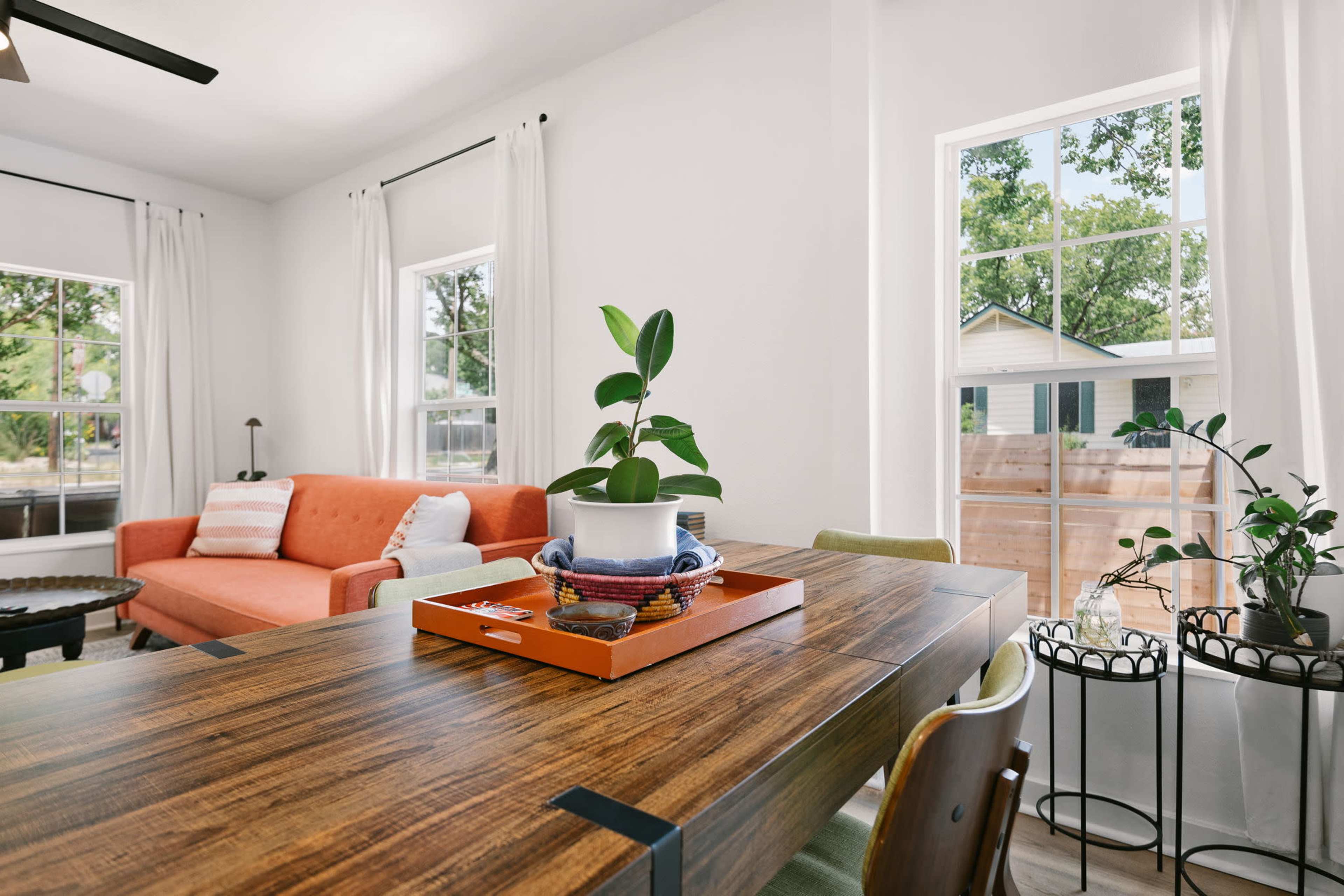 A bright living area features a wooden dining table with a decorative tray, an orange sofa, and large windows allowing natural light to fill the space.