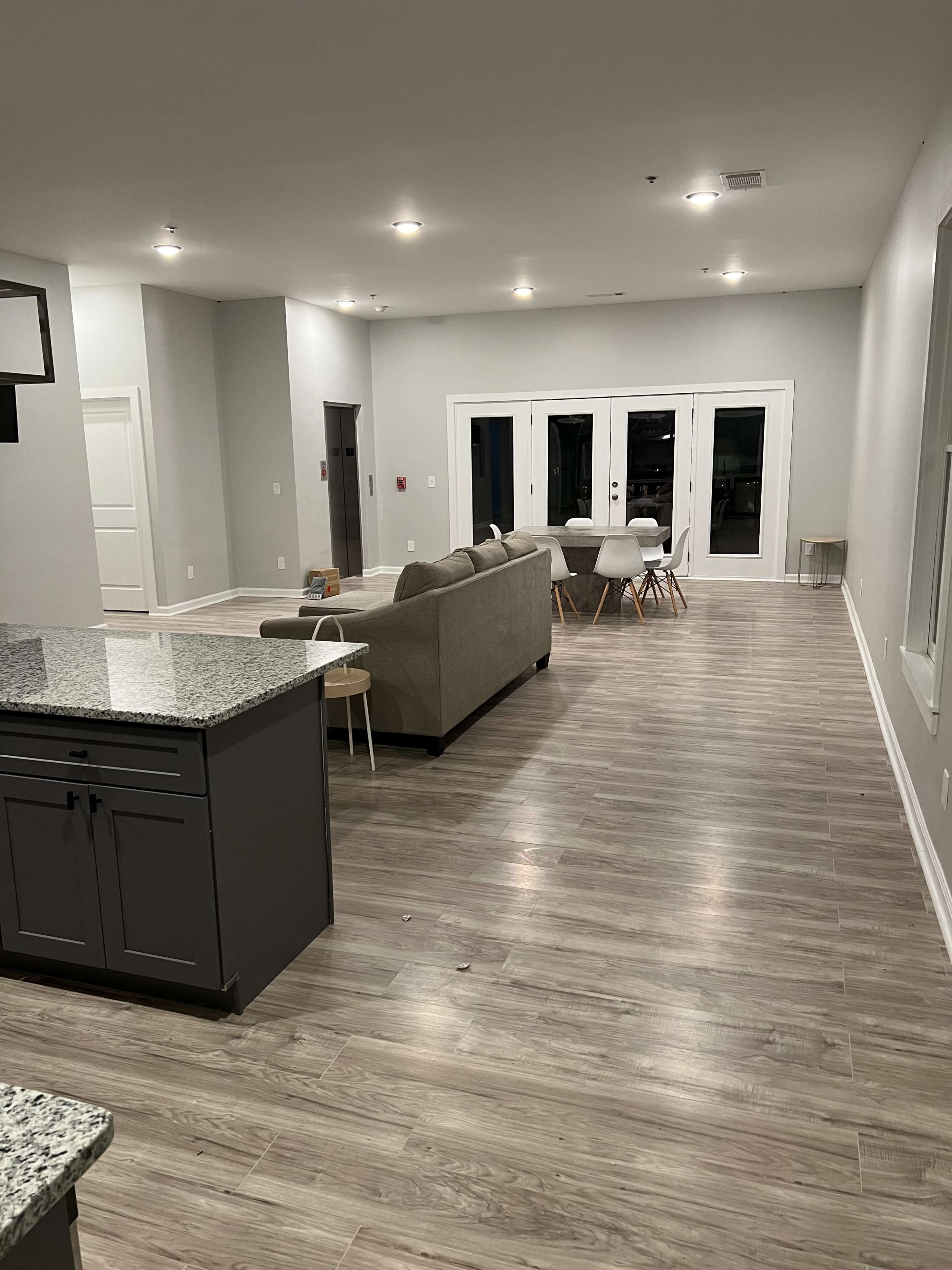 The image shows a spacious, modern living area with light-colored walls, hardwood flooring, a gray sofa, and a dining area with a table and chairs in the background.