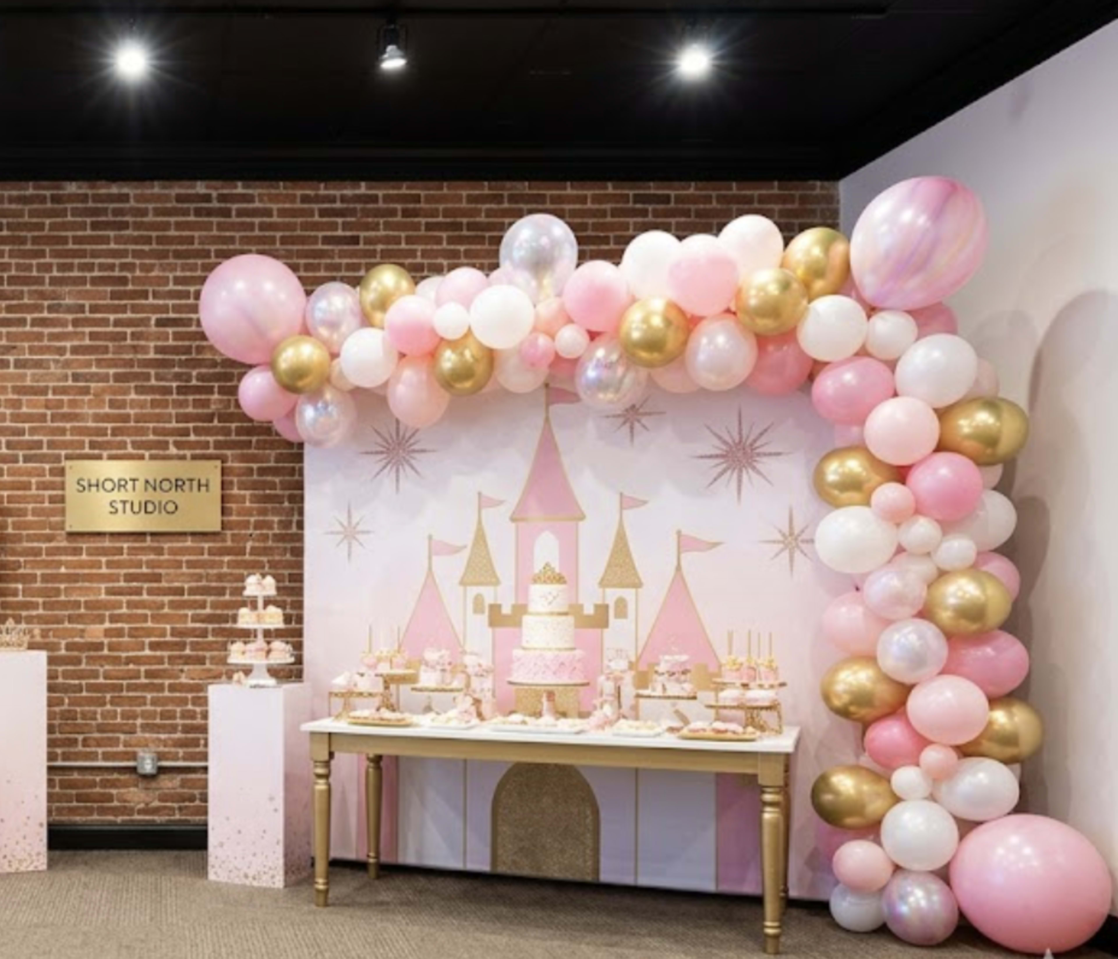 The image shows a decorative party setup featuring a table with a dessert display, a large cake, and an arch made of pink, white, and gold balloons, all set against a brick wall and a themed backdrop.