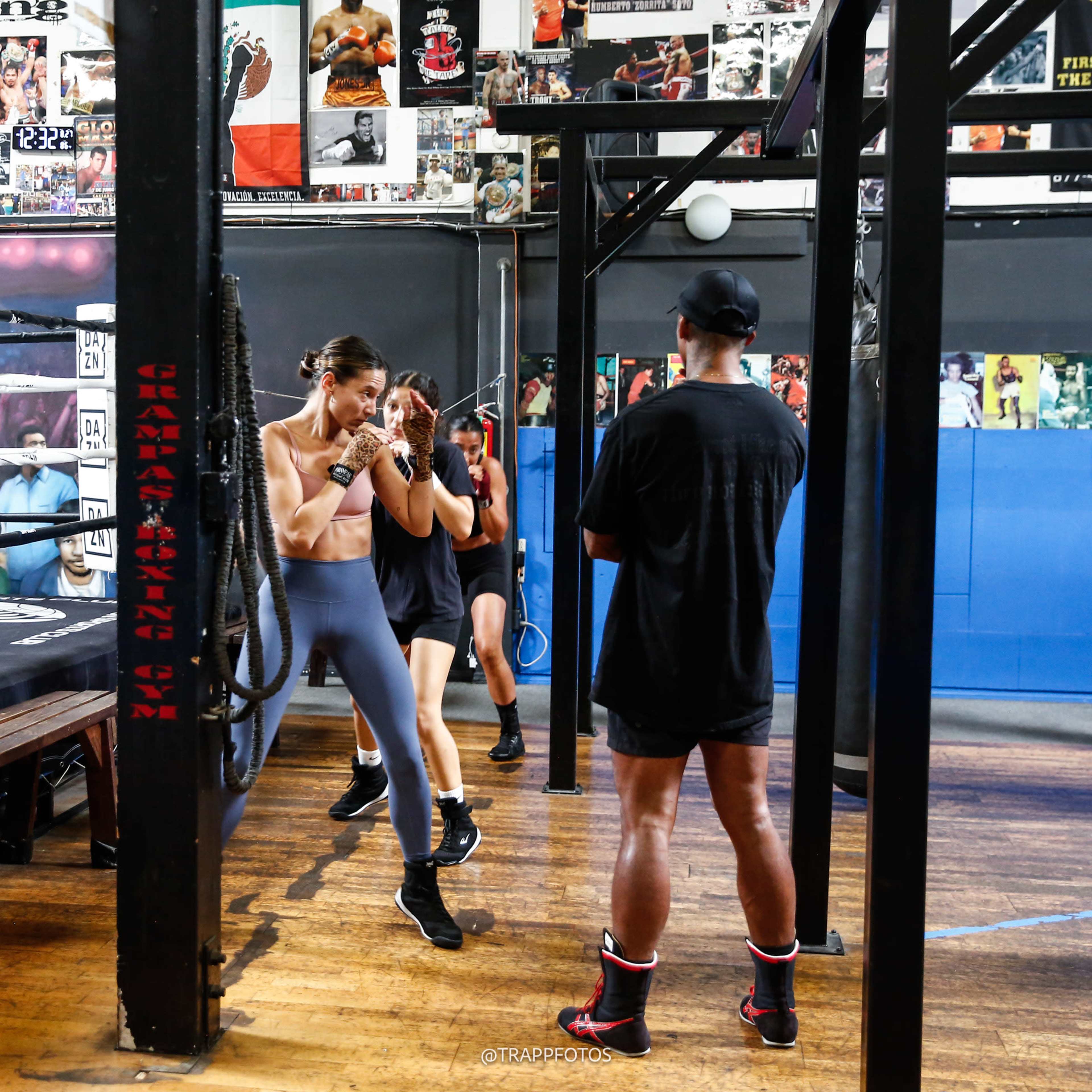 Three individuals train in a boxing gym, with one person practicing punches on a heavy bag while two others observe.