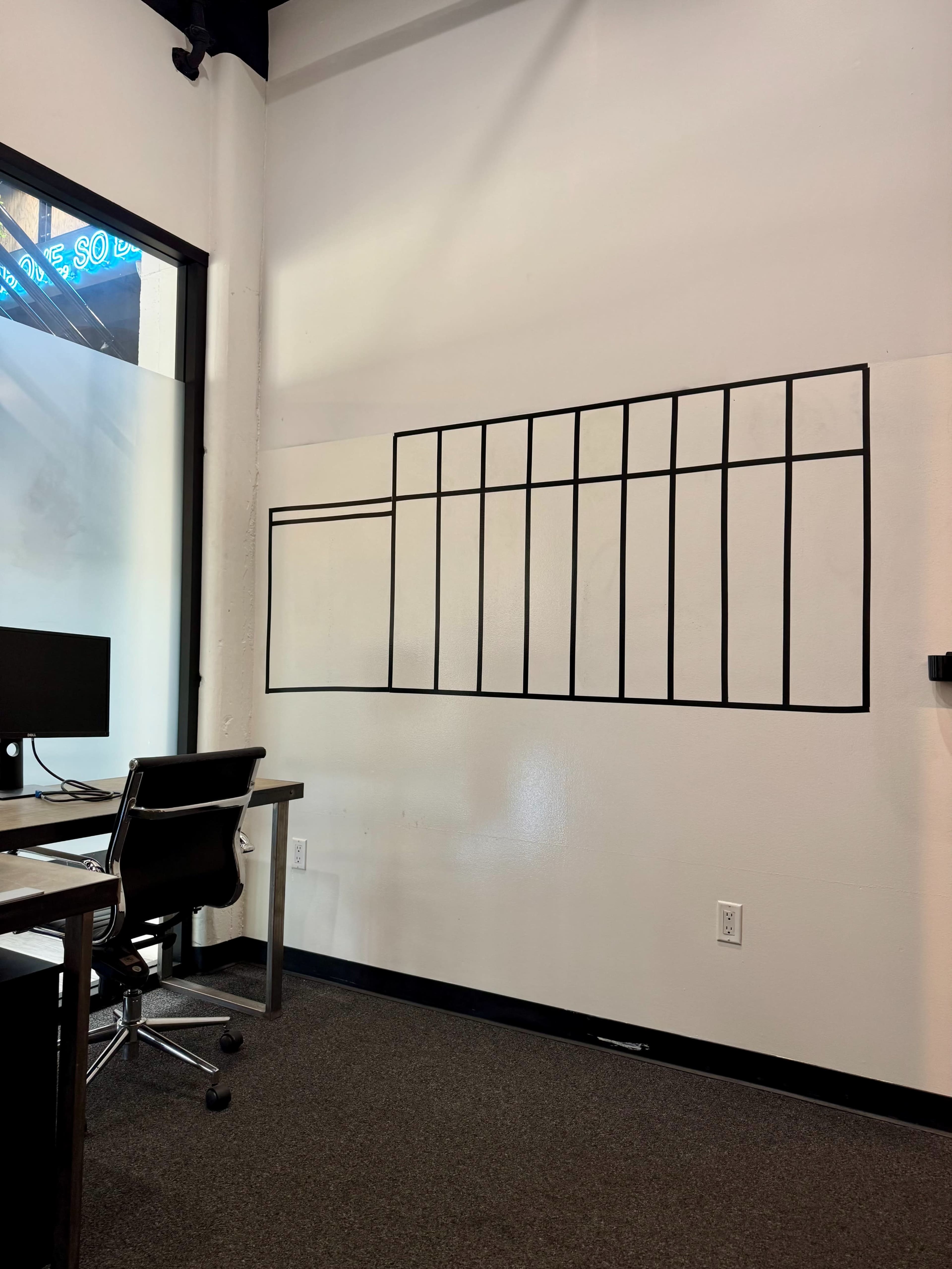 A wall in an office features a grid of black tape arranged in a rectangular pattern, alongside a desk with an office chair.