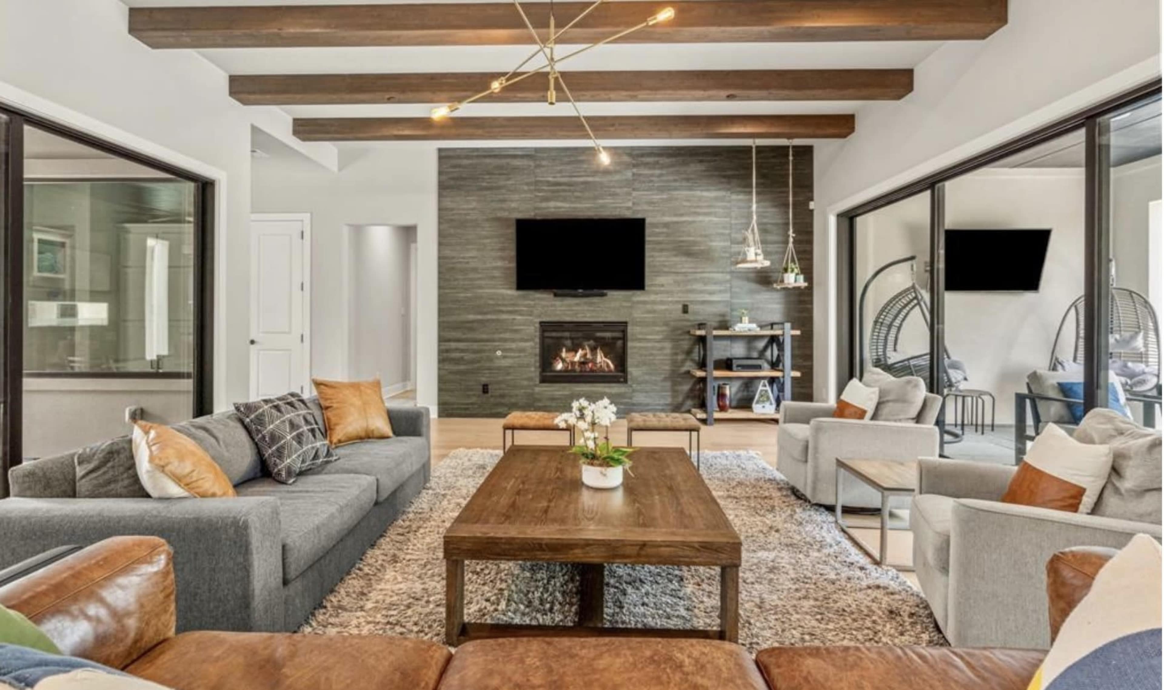 The living room features a combination of gray and brown furniture arranged around a large wooden coffee table, with a fireplace and a mounted TV on the stone accent wall.