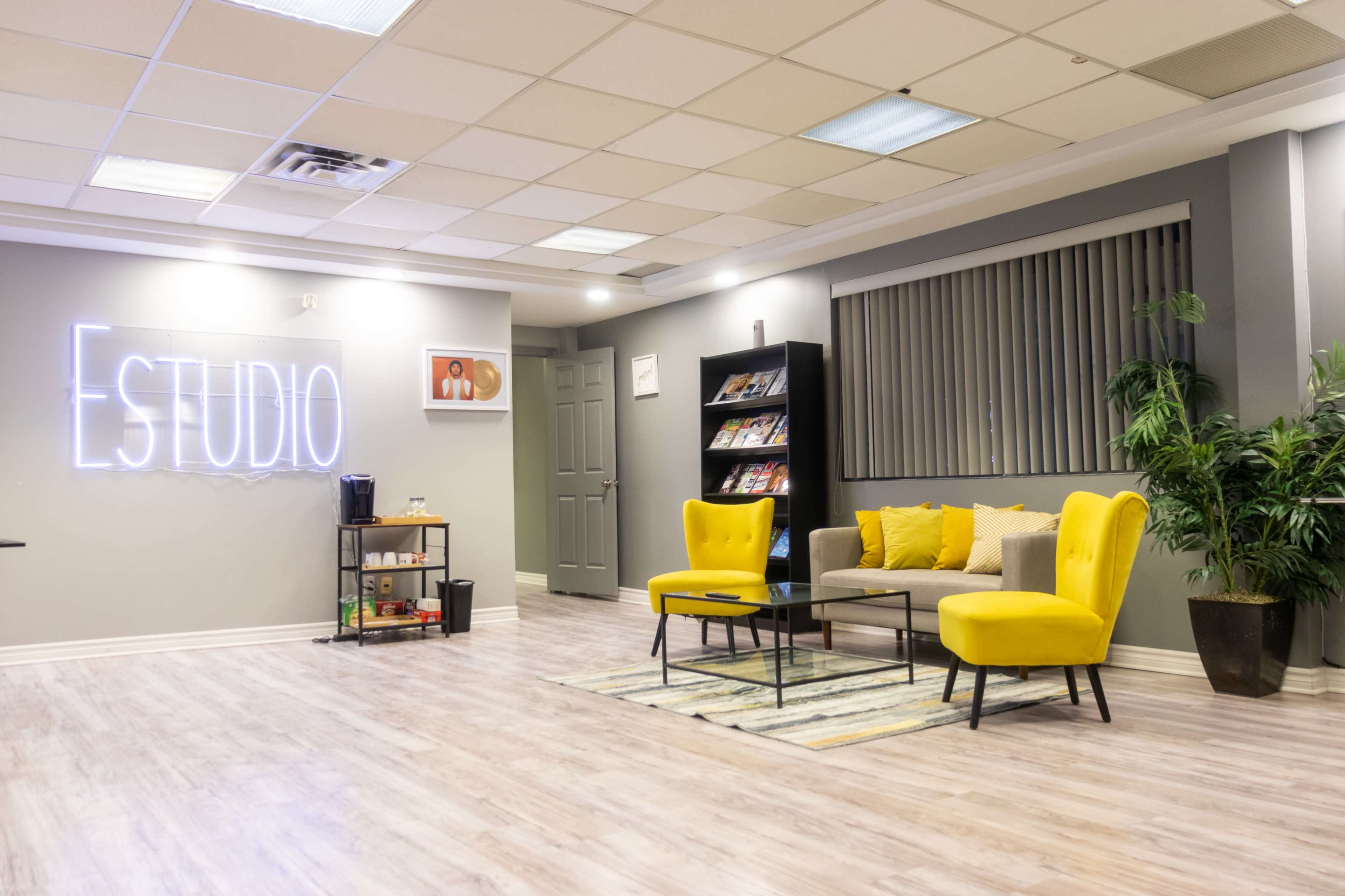 A modern studio space with neon signage reading "ESTUDIO," two yellow chairs, a gray sofa, and a bookshelf filled with magazines.