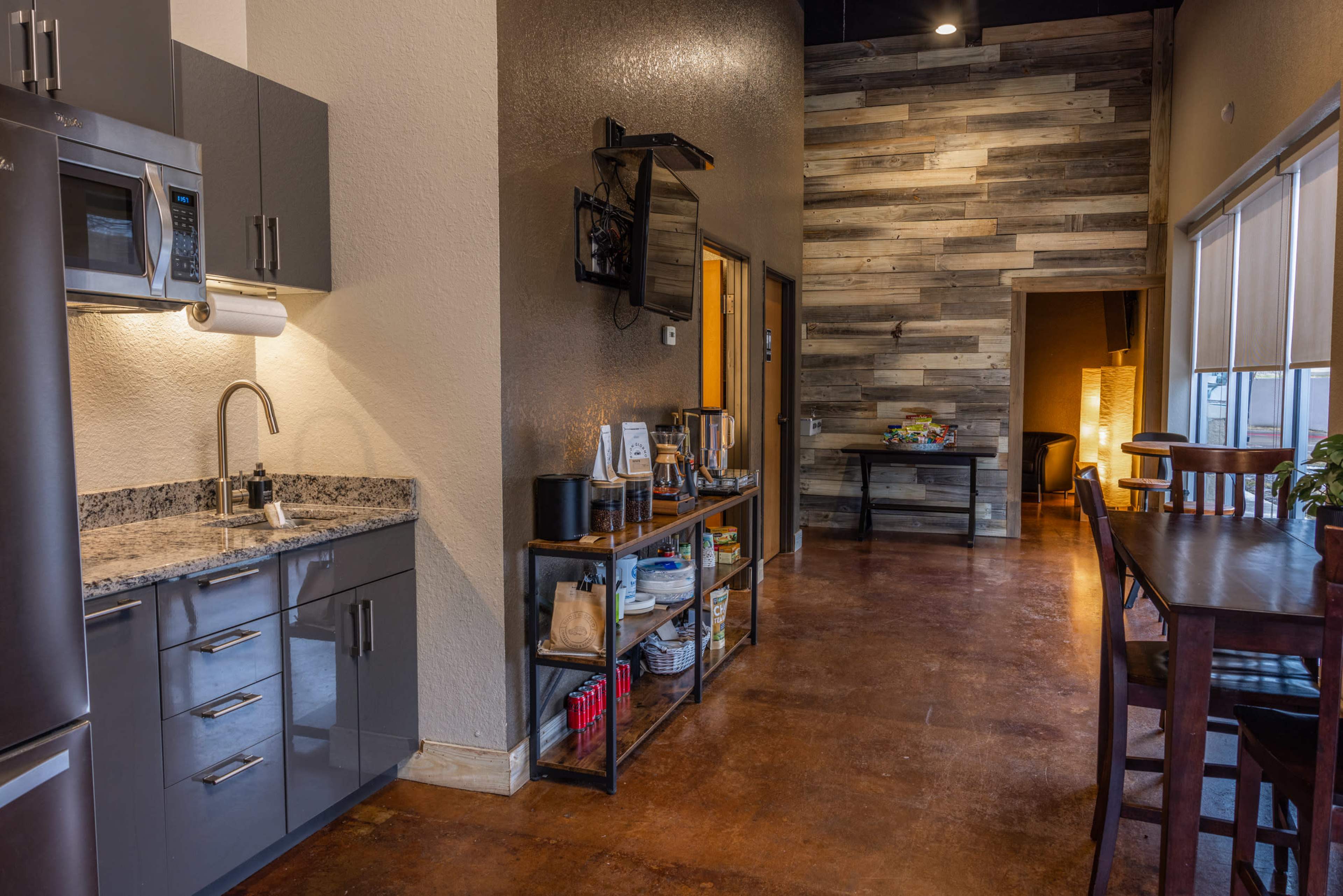 A modern kitchen area features stainless steel appliances and a shelf with various items, adjacent to a wooden-paneled wall and a dining area.