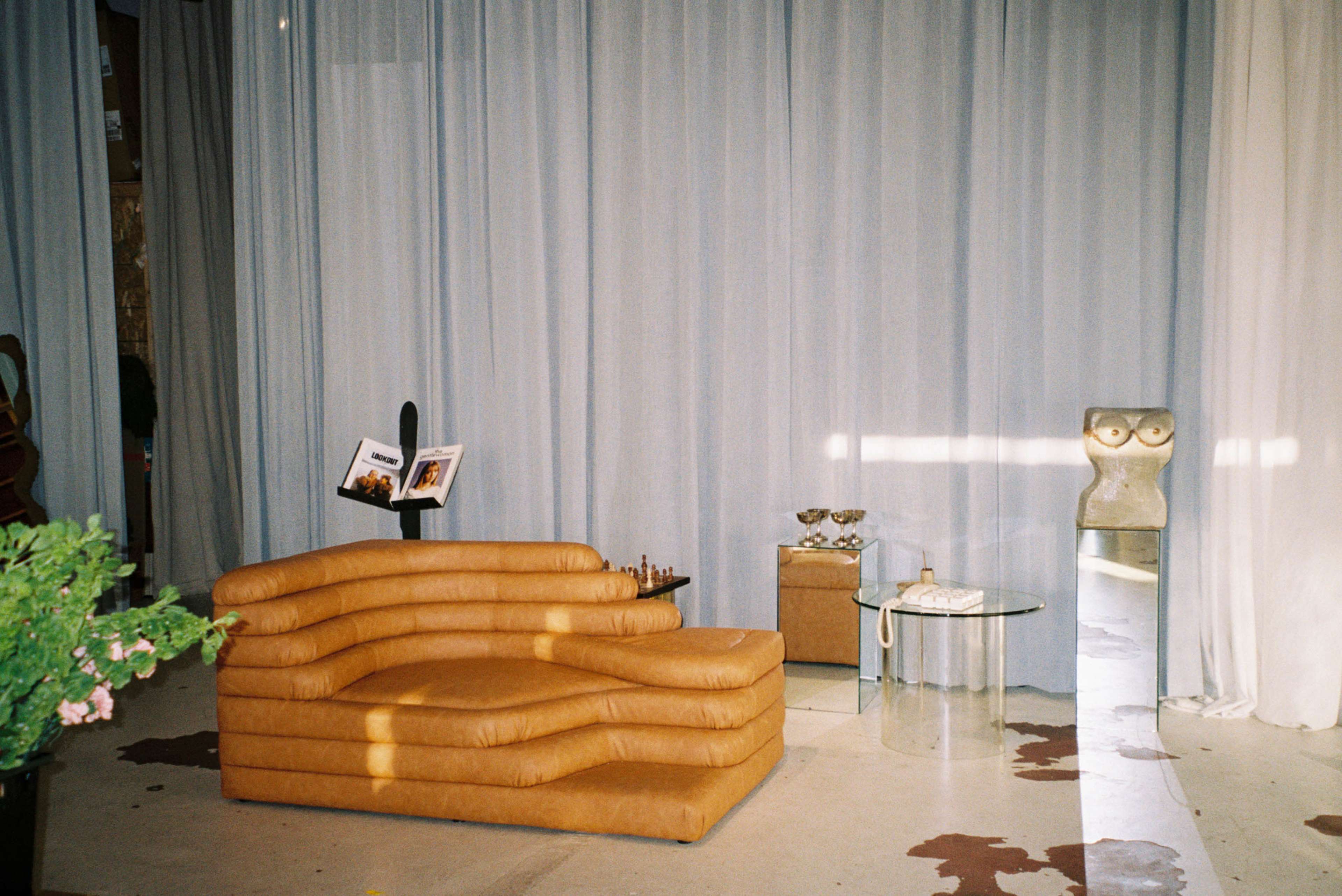 A tan, curved sofa is positioned in a room with sheer curtains, accompanied by a clear glass table and a sculptural figure.