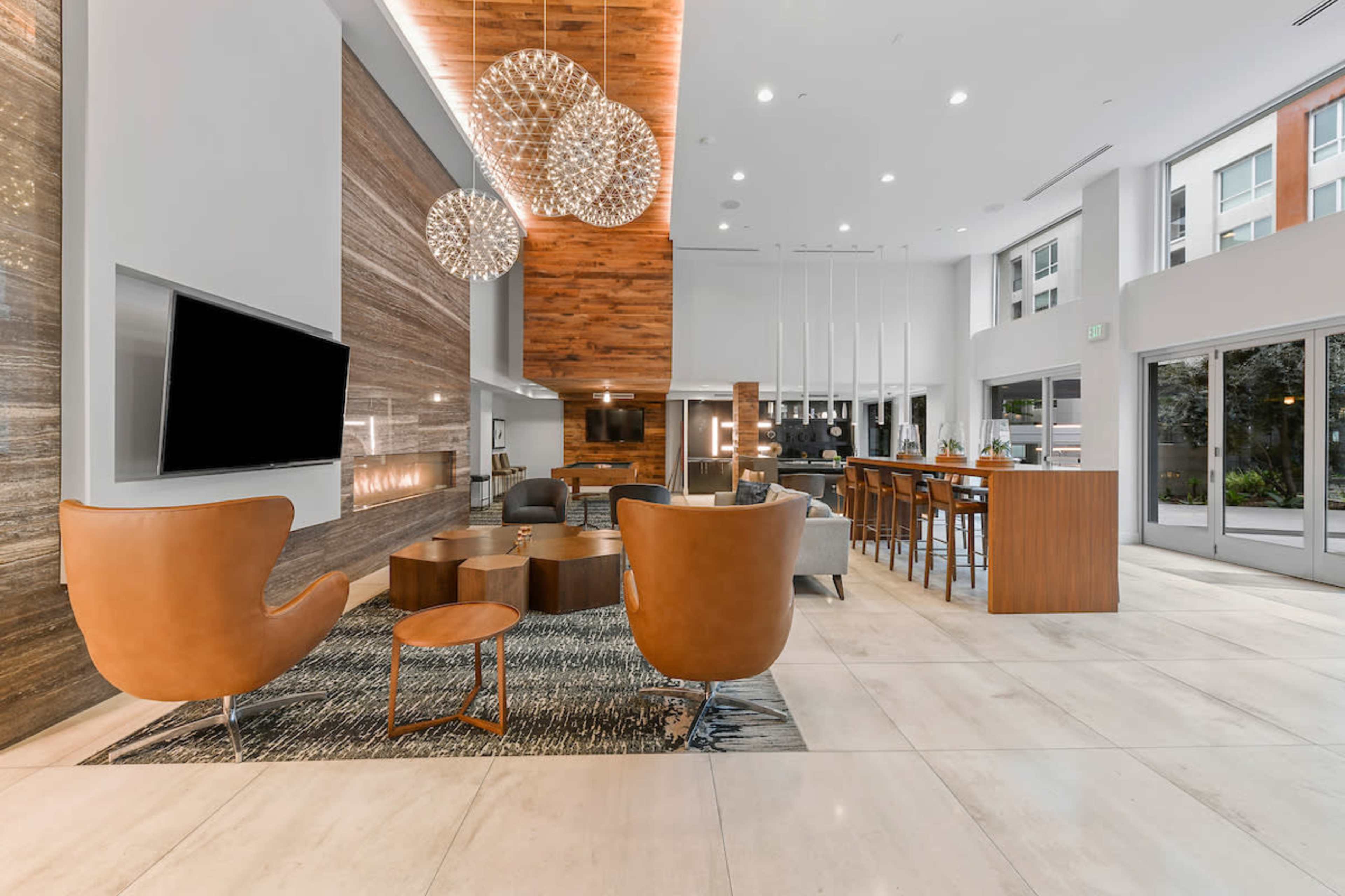 A modern lobby with a seating area, a television, decorative lighting fixtures, and a bar-style dining table adjacent to large windows.
