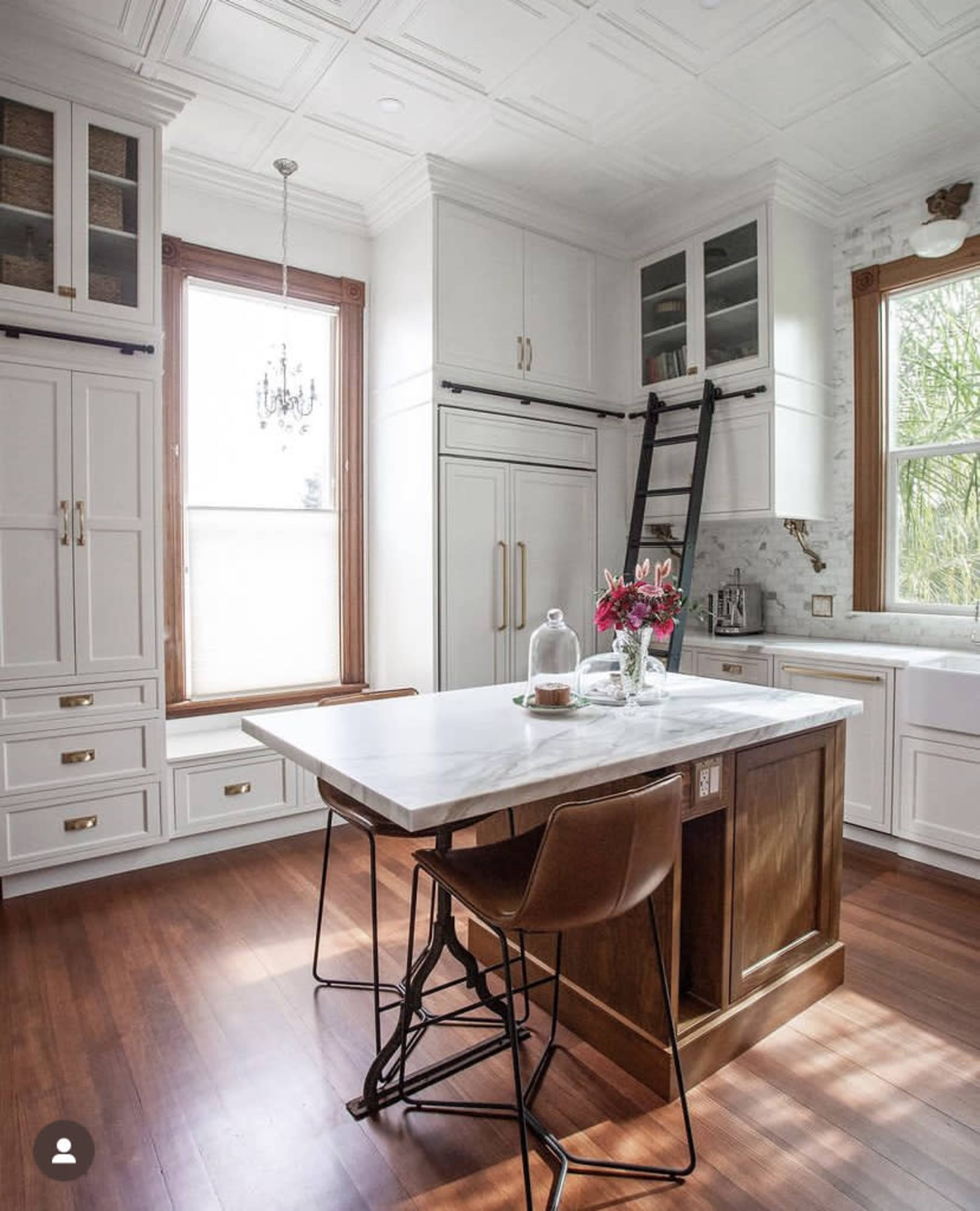 A modern kitchen featuring white cabinetry, a marble-topped island, and a wooden ladder against a wall, with natural light streaming in through large windows.