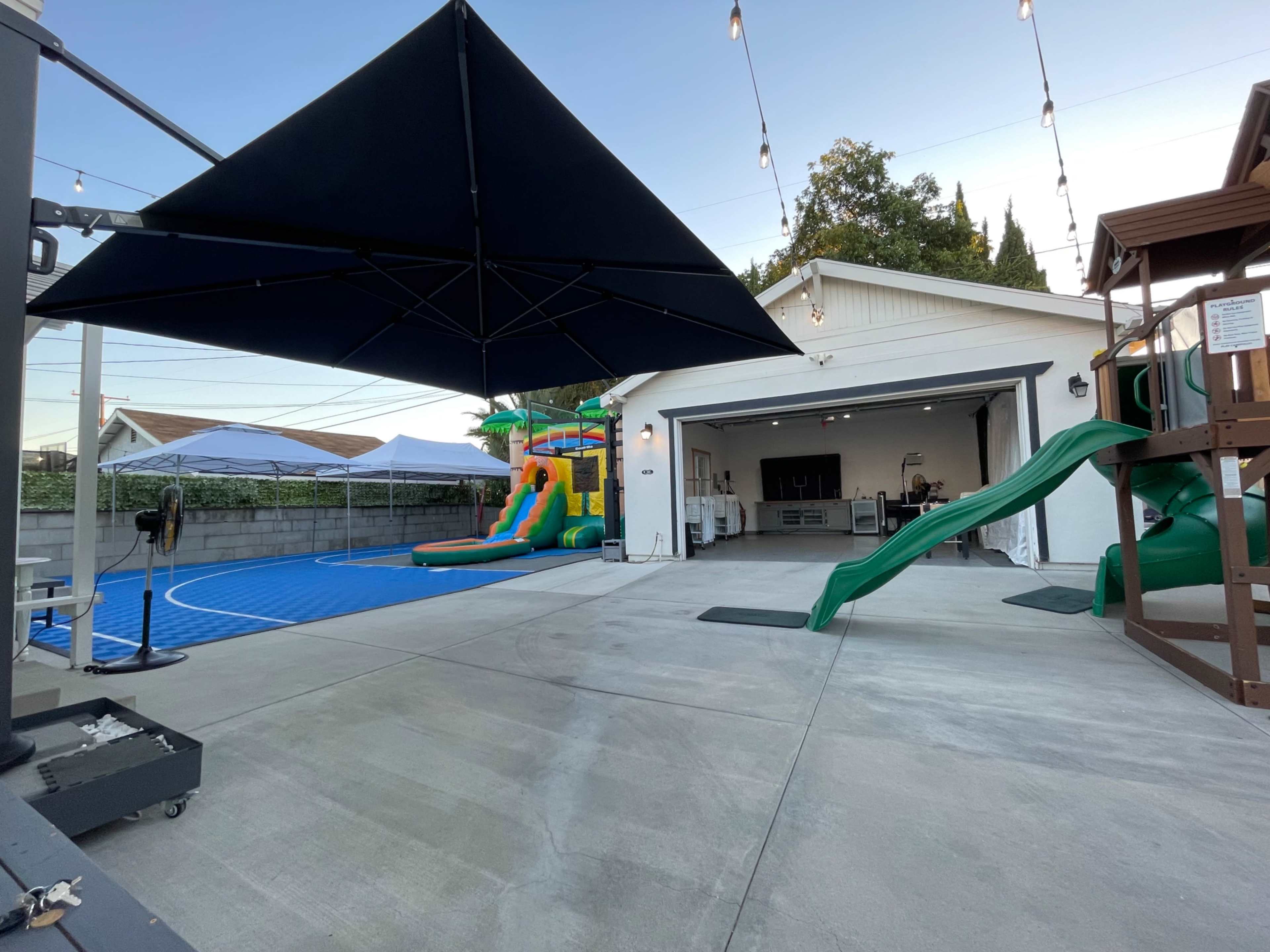 The image shows a backyard area featuring a large swimming pool with a slide, a colorful inflatable play structure, and a covered outdoor seating space.
