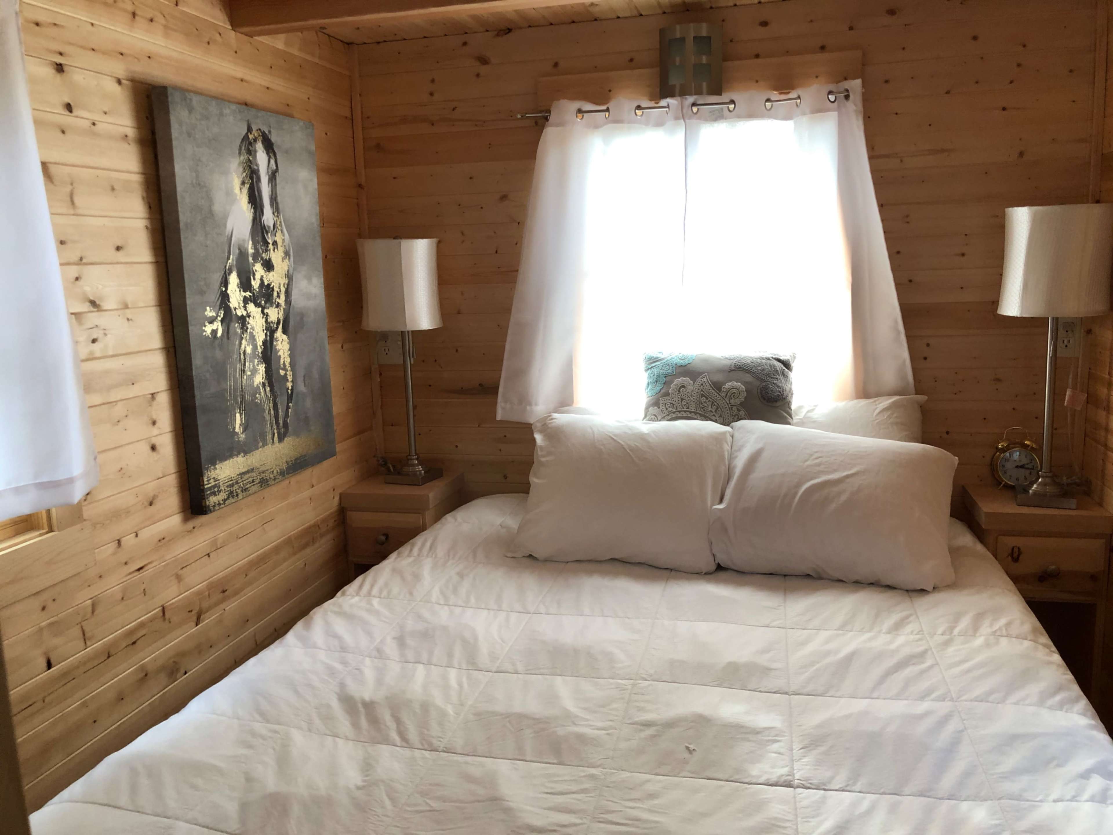A cozy bedroom with a wooden interior, featuring a bed with white pillows and a blanket, flanked by two lamps and a wall art depicting a horse.