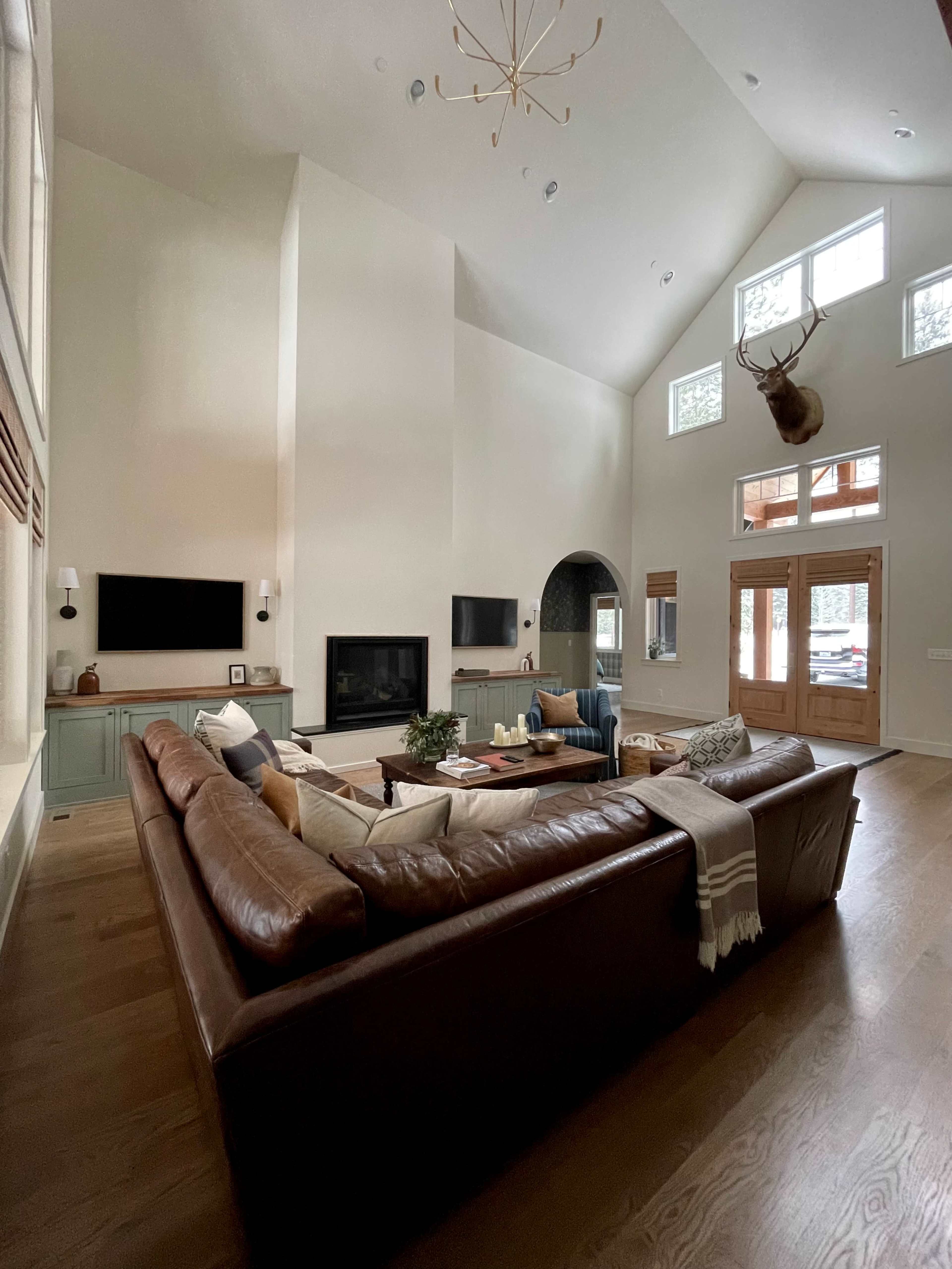 A spacious living room features a large leather sectional sofa, two wall-mounted televisions, and tall windows that fill the space with light.