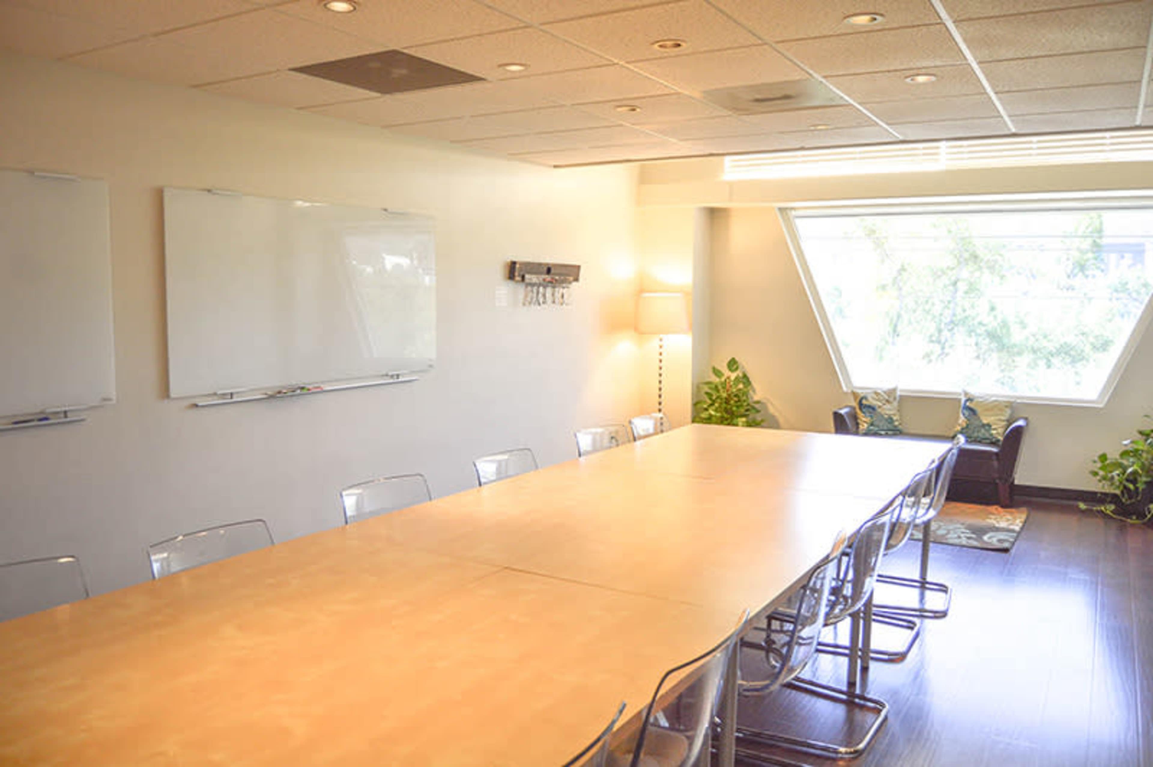 A spacious conference room features a long table, clear walls for presentations, and a large window overlooking greenery.