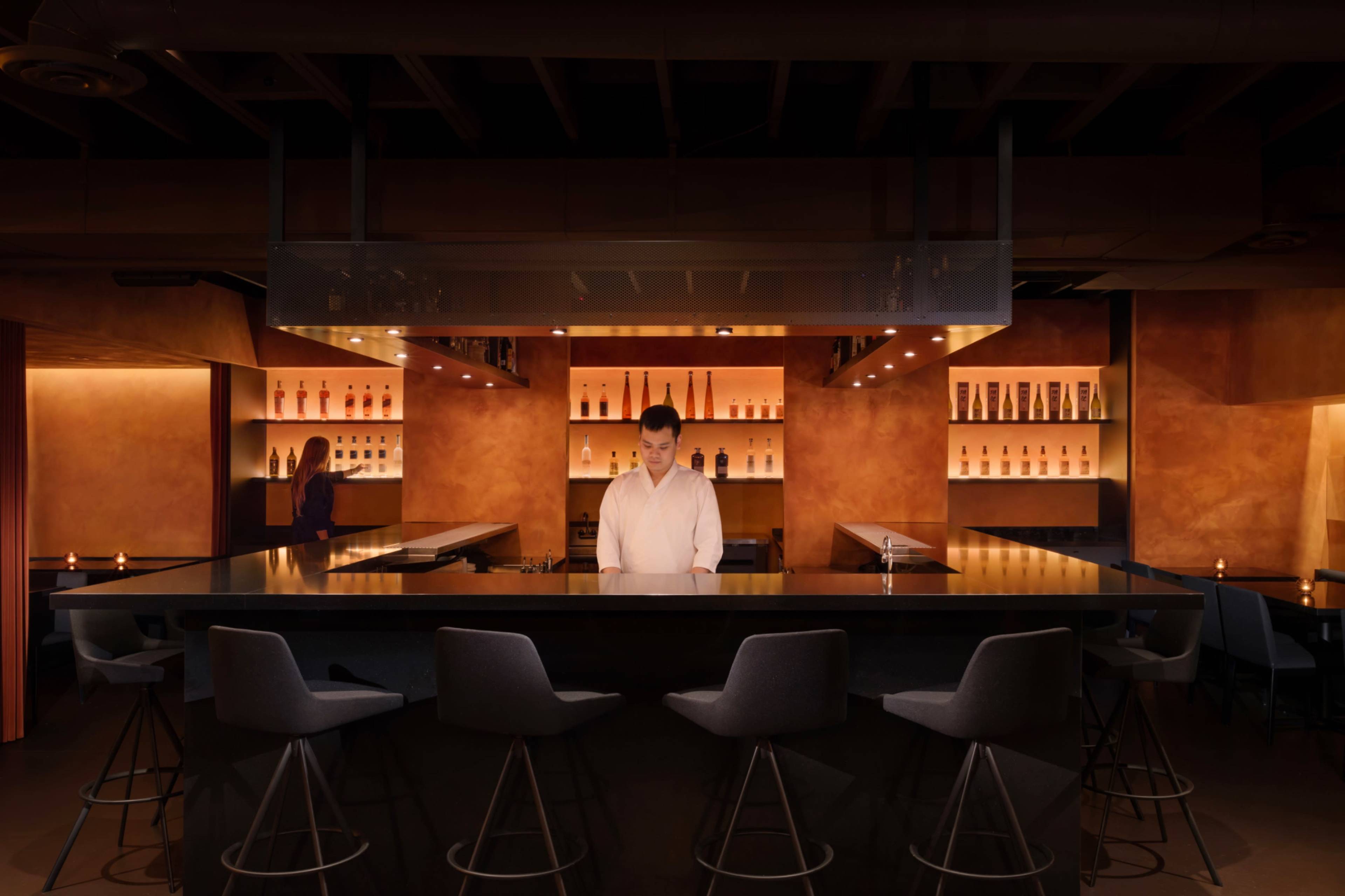 A bartender stands alone at a dimly lit bar with illuminated bottles displayed on shelves behind him.