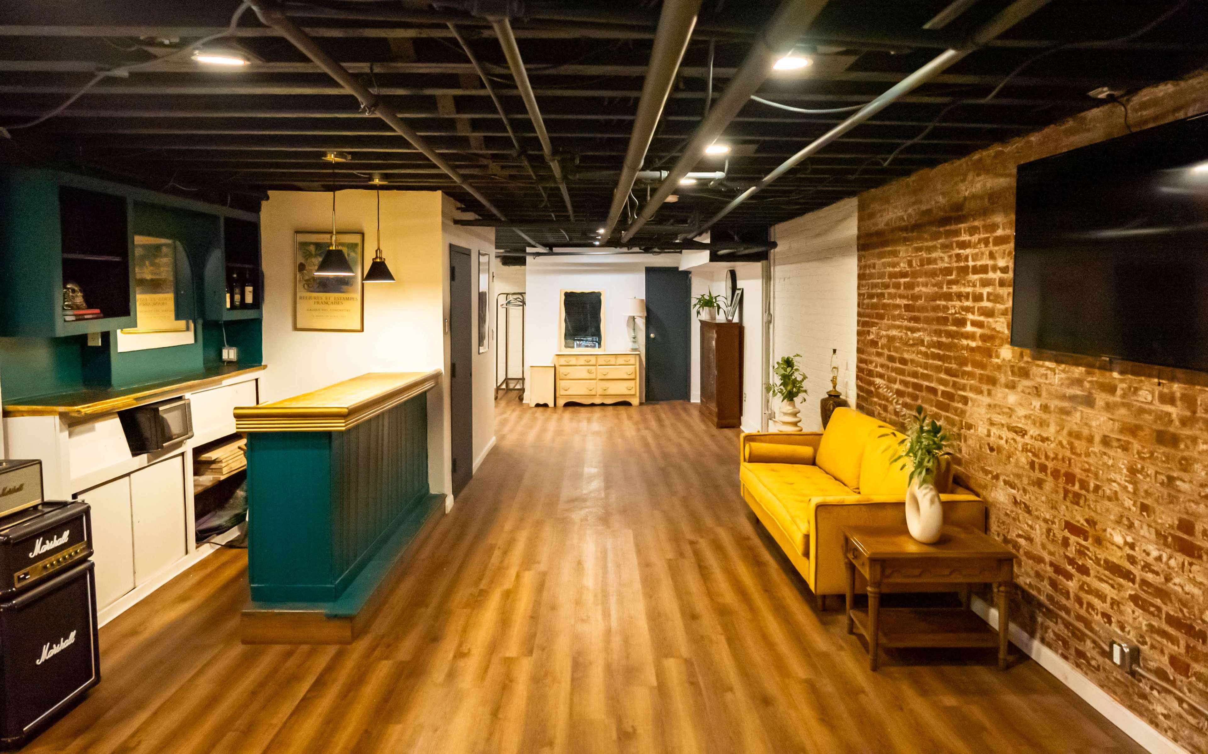 A modern interior space featuring a long, narrow hallway with wooden flooring, a yellow sofa, exposed brick walls, and a teal accent wall with shelves.