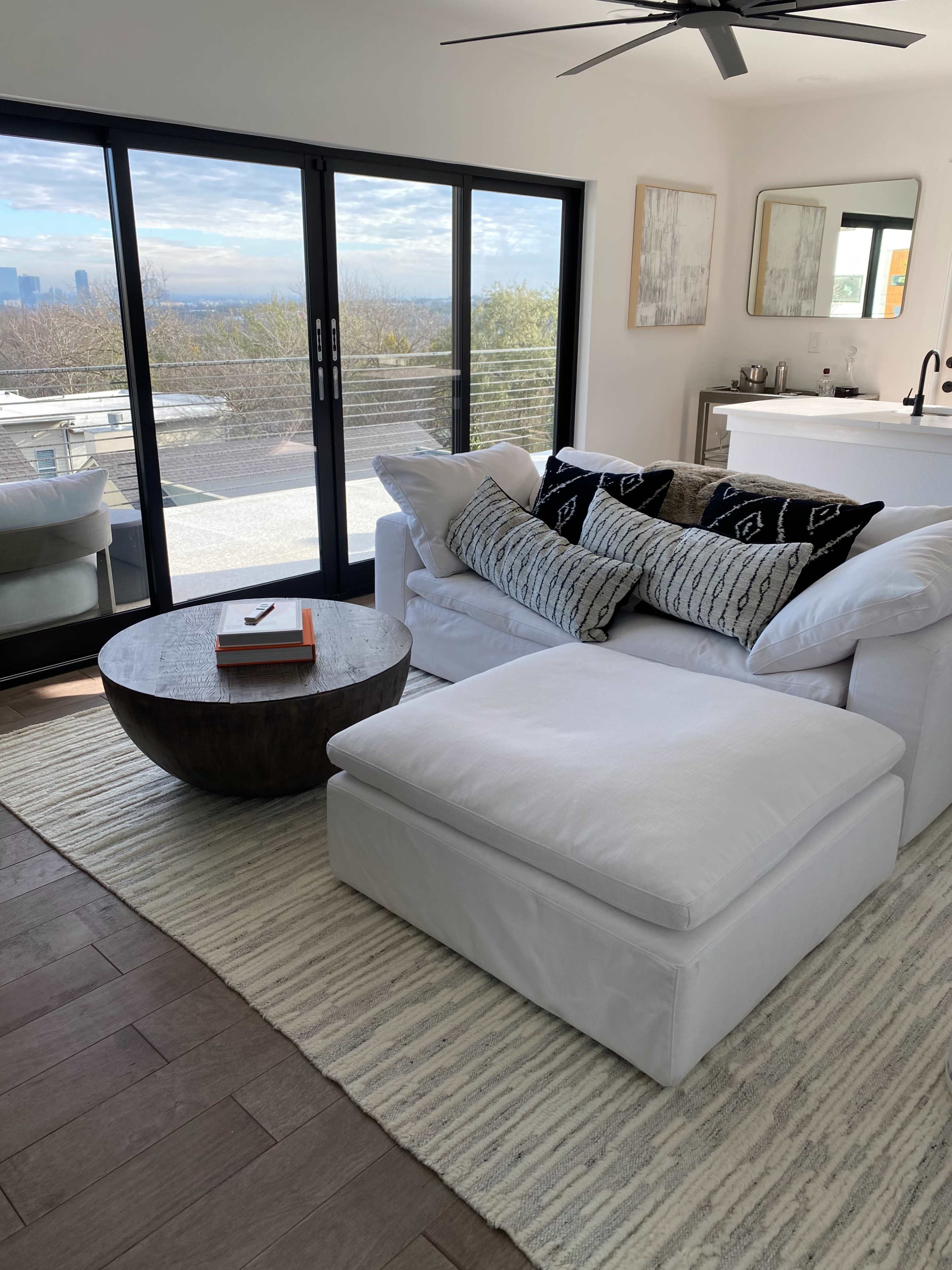 A bright living room features a white sectional sofa with black and white patterned pillows, a round wooden coffee table, and a large window offering a view of the city skyline.