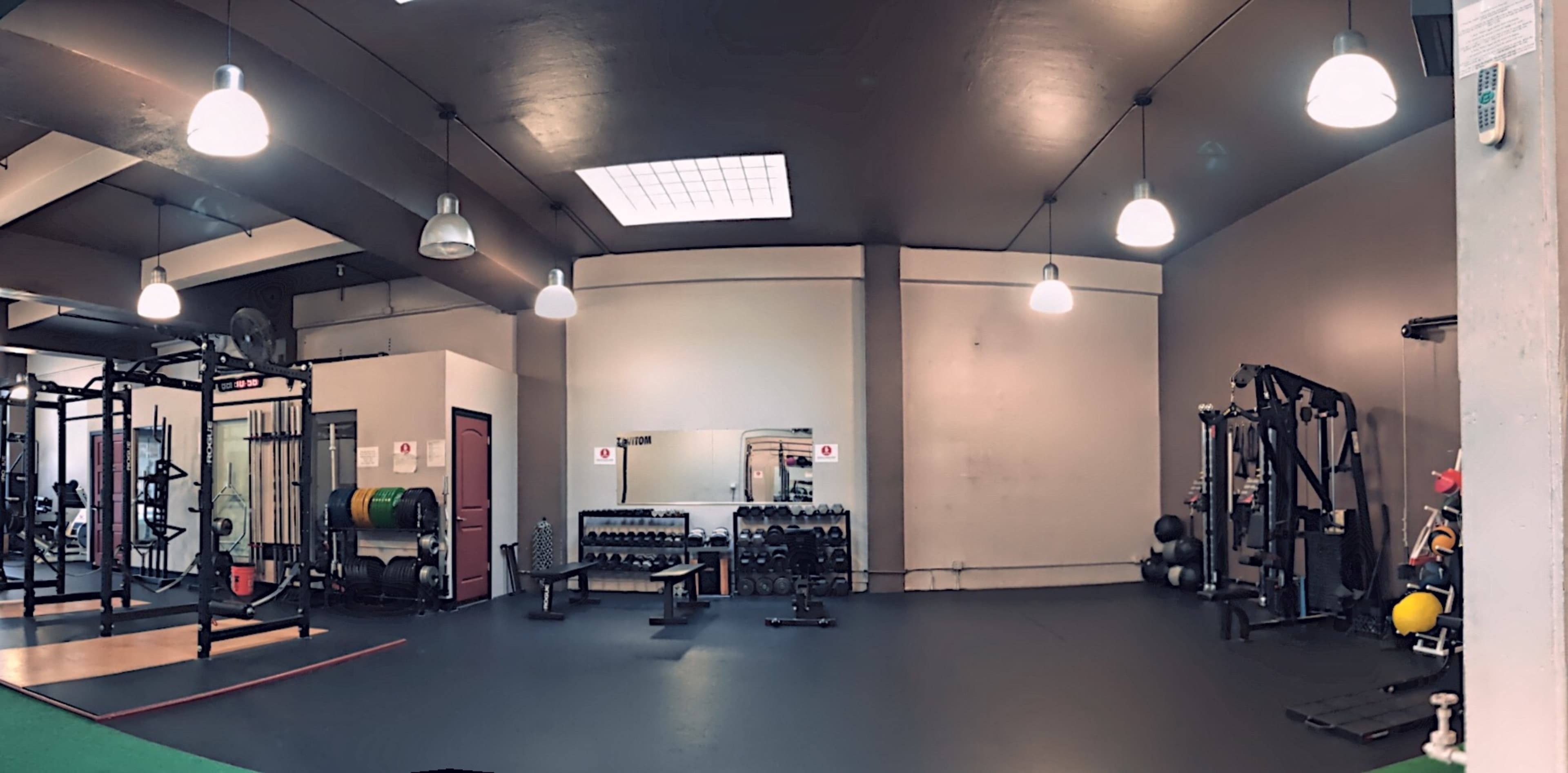 A spacious gym interior featuring various workout equipment, including weight racks, cable machines, and free weights, all under bright ceiling lights.