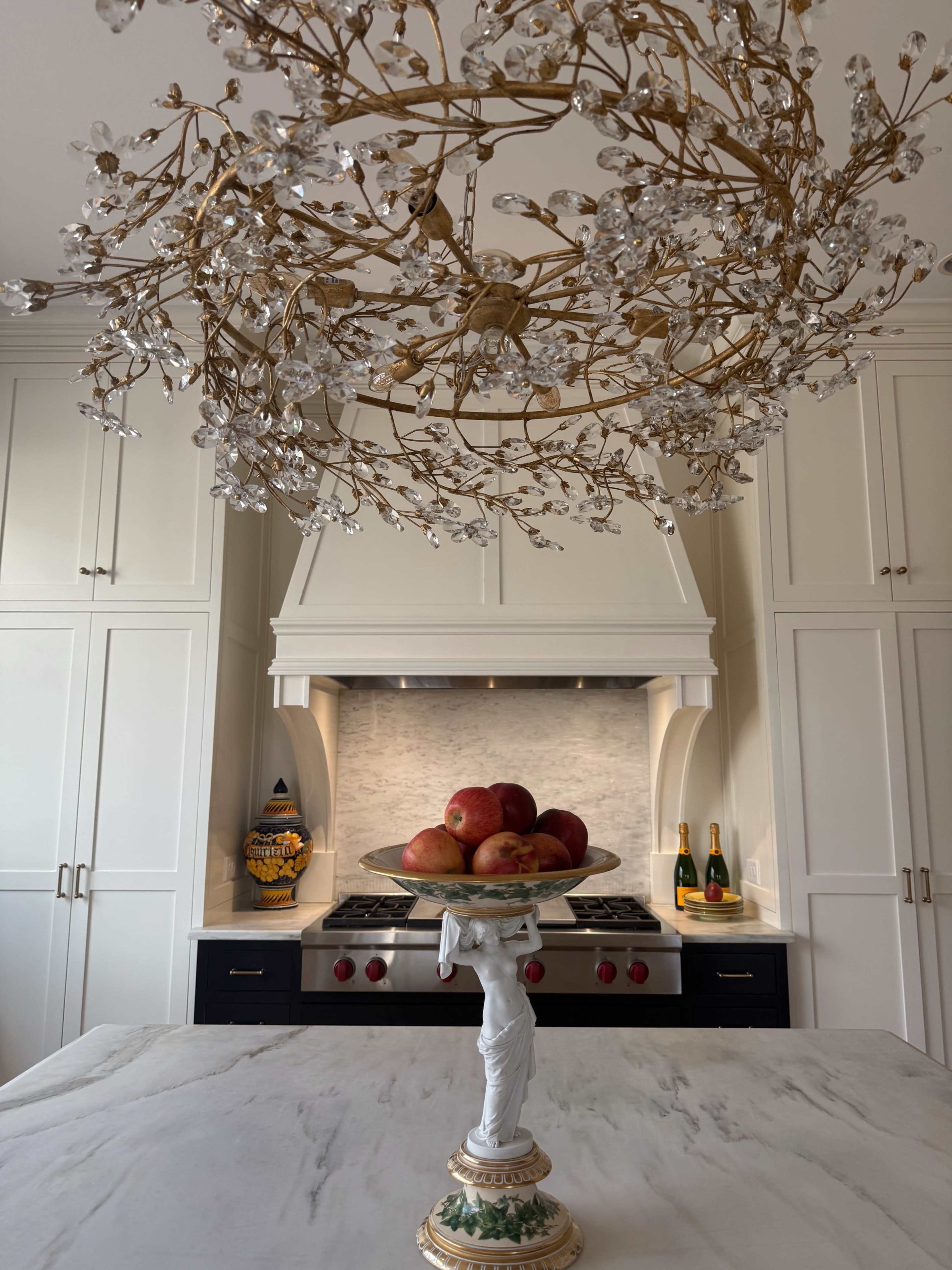 A kitchen features a chandelier made of crystal and gold accents, with a marble island displaying a pedestal bowl of red apples in front of a stove and cabinetry.