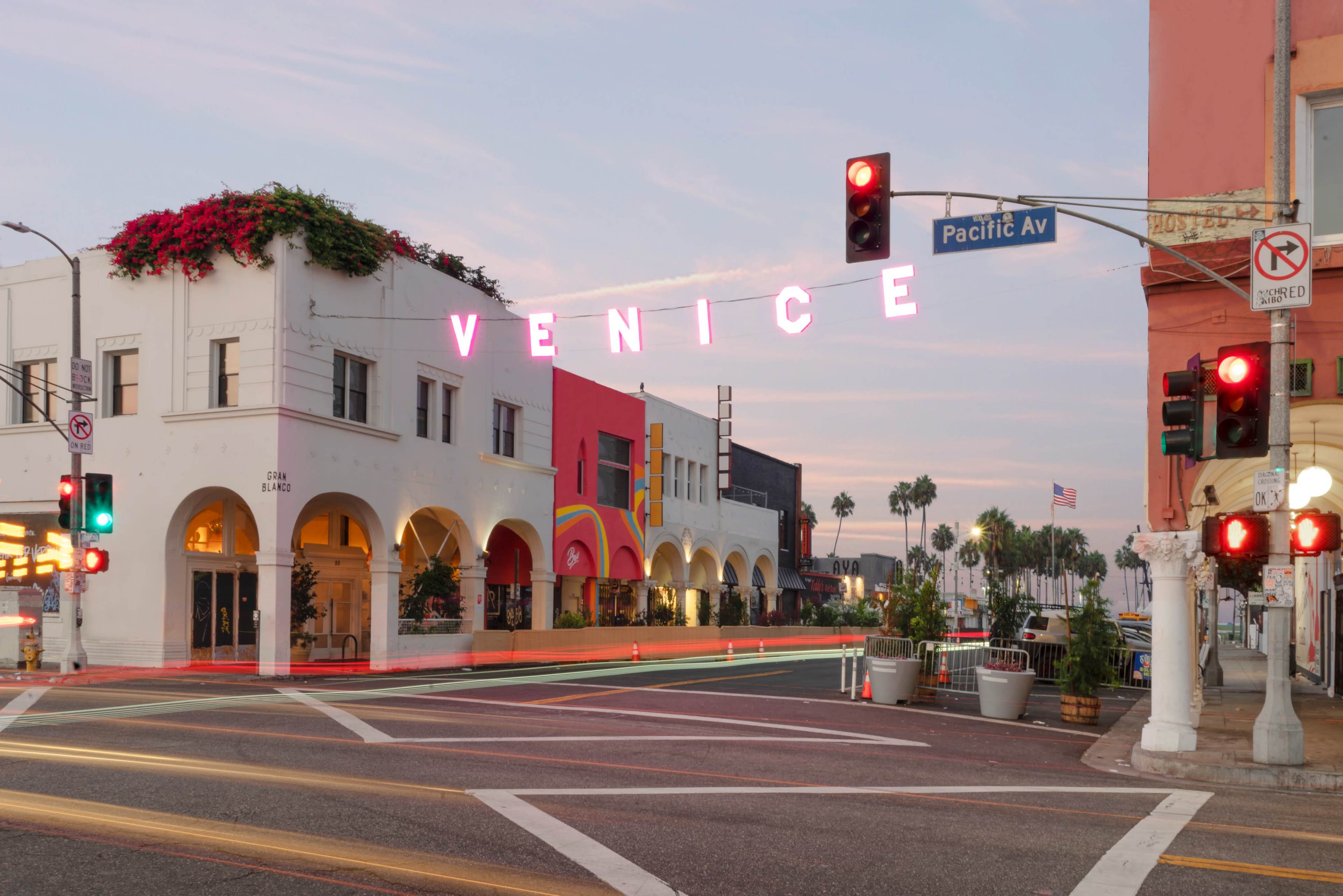The image shows a street scene in Venice, California, featuring buildings with vibrant facades and a neon sign that reads "VENICE" above the intersection at Pacific Avenue.