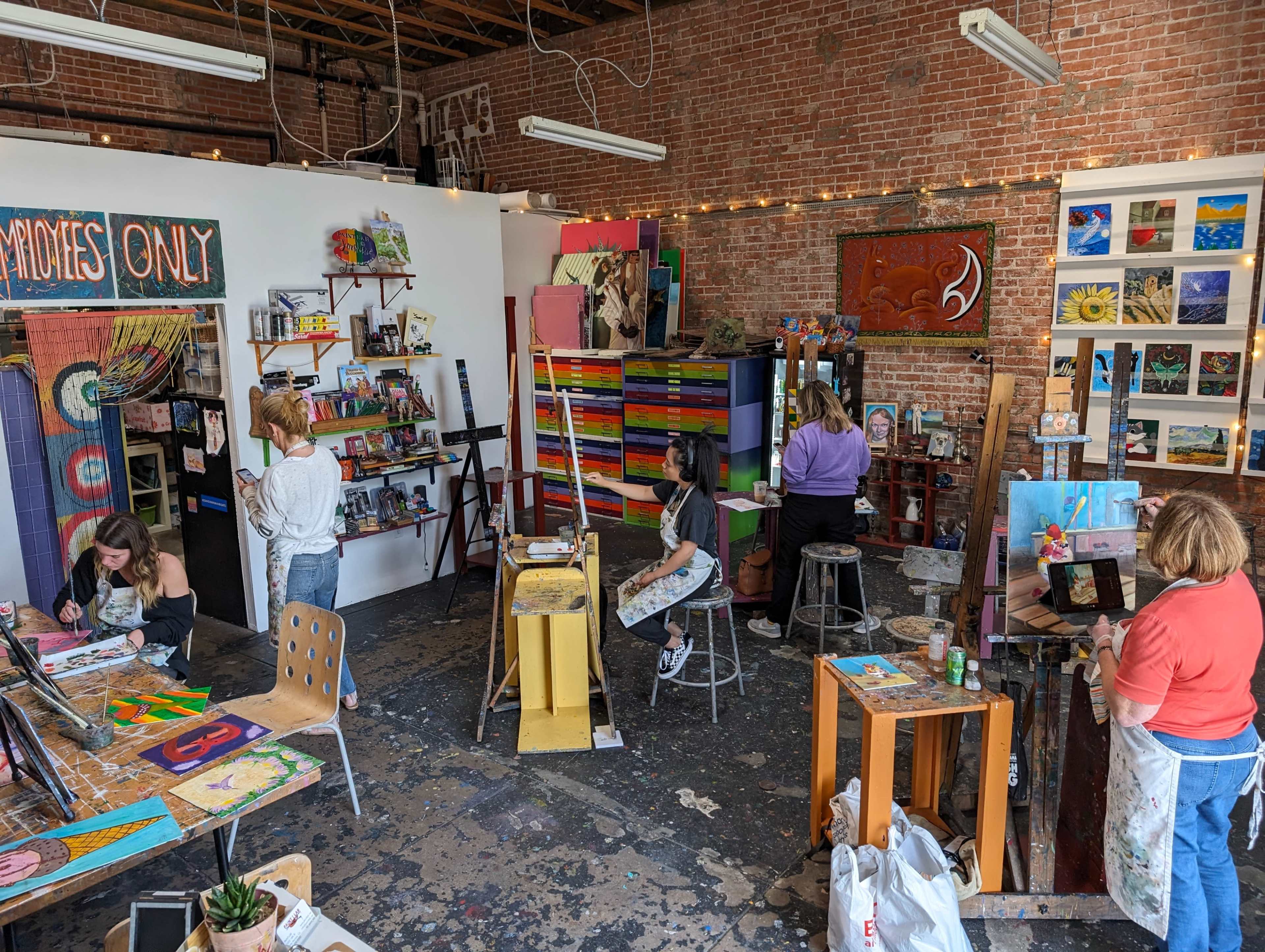 The image shows an art studio with several people engaged in painting, surrounded by colorful artwork and shelves filled with art supplies.