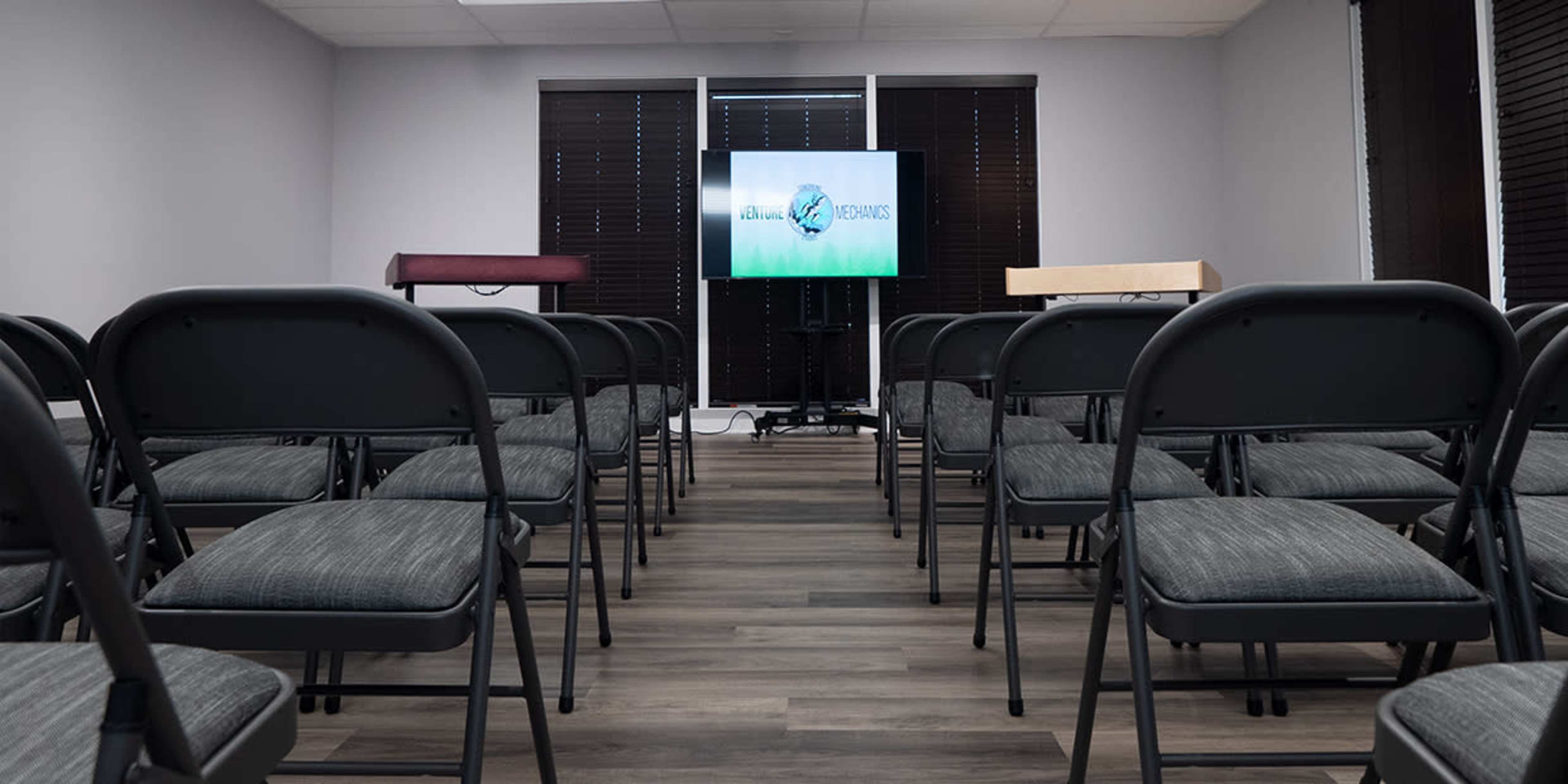 A meeting room with rows of folding chairs facing a screen displaying the logo of a company or organization.