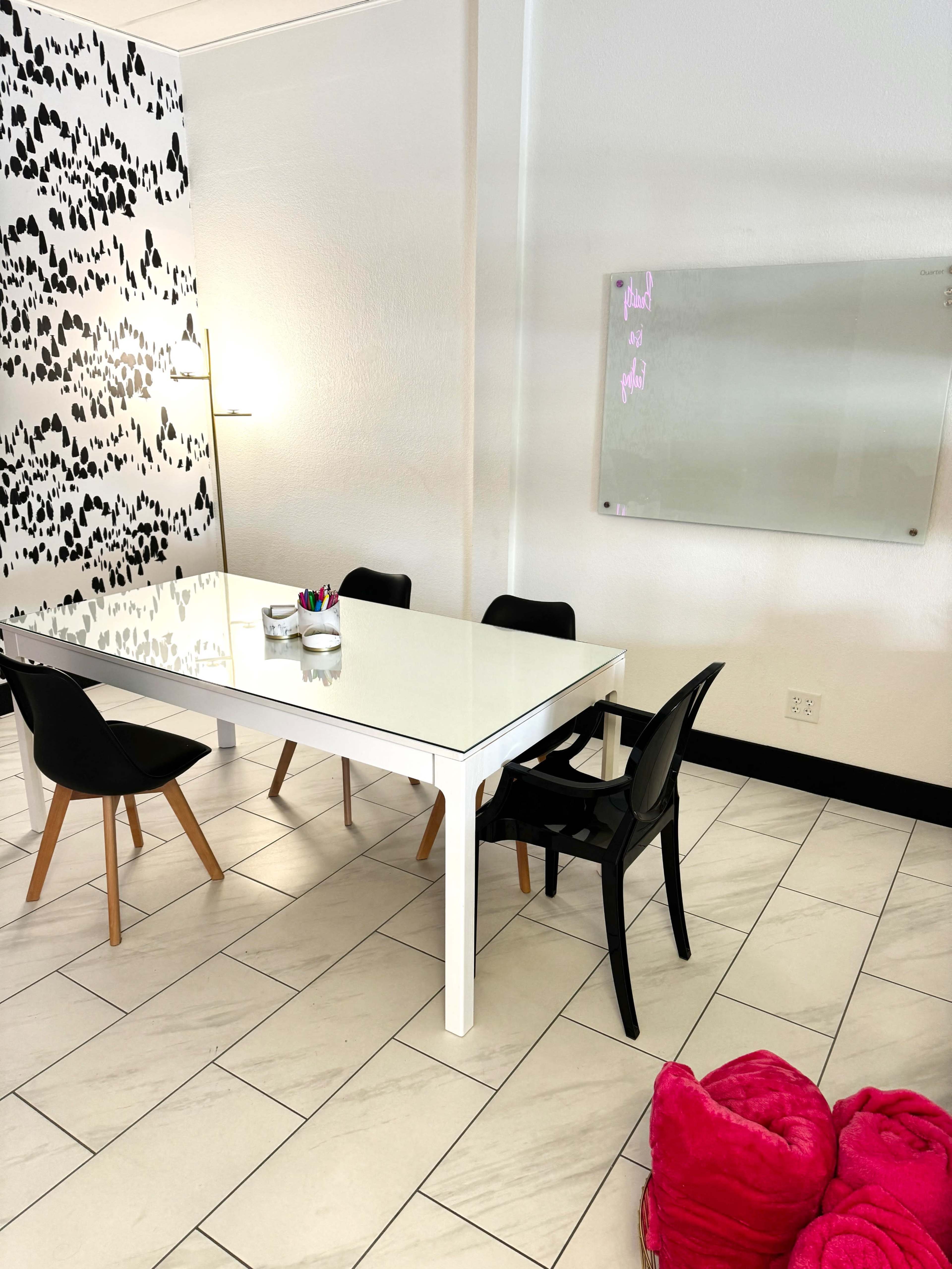A modern meeting room features a white table surrounded by black chairs, with a patterned wall and a glass whiteboard.