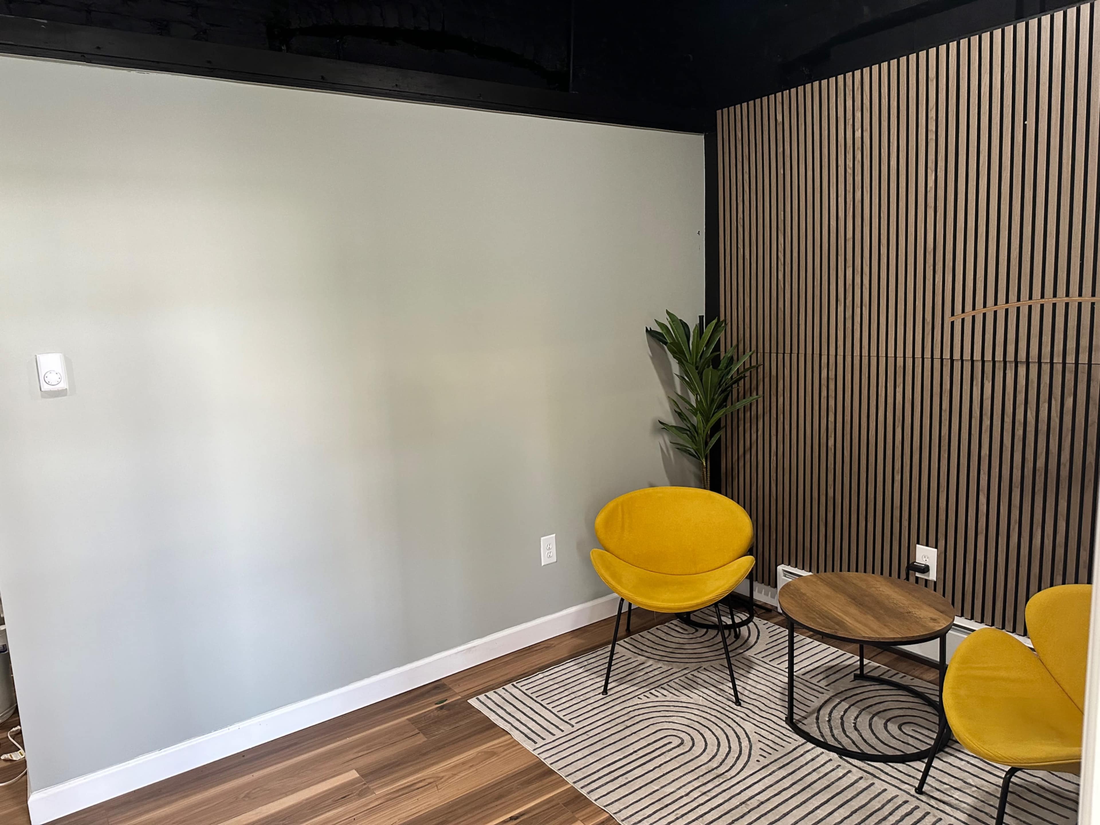 The image shows a small seating area featuring two yellow chairs, a round table, a potted plant, and a wall with vertical wooden panels.