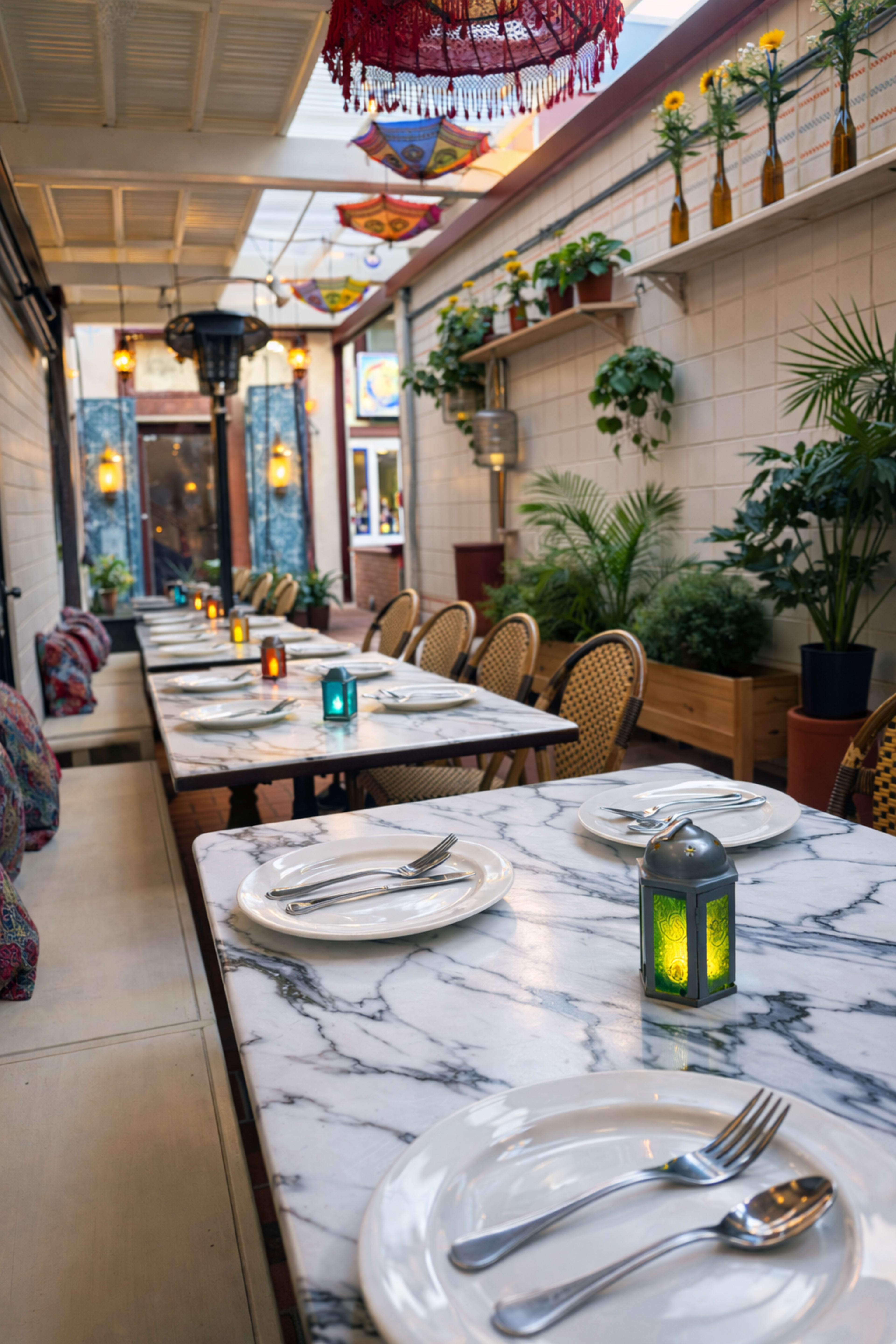 The image shows a decorated indoor dining area with marble tables set for meals, surrounded by plants and colorful lanterns.