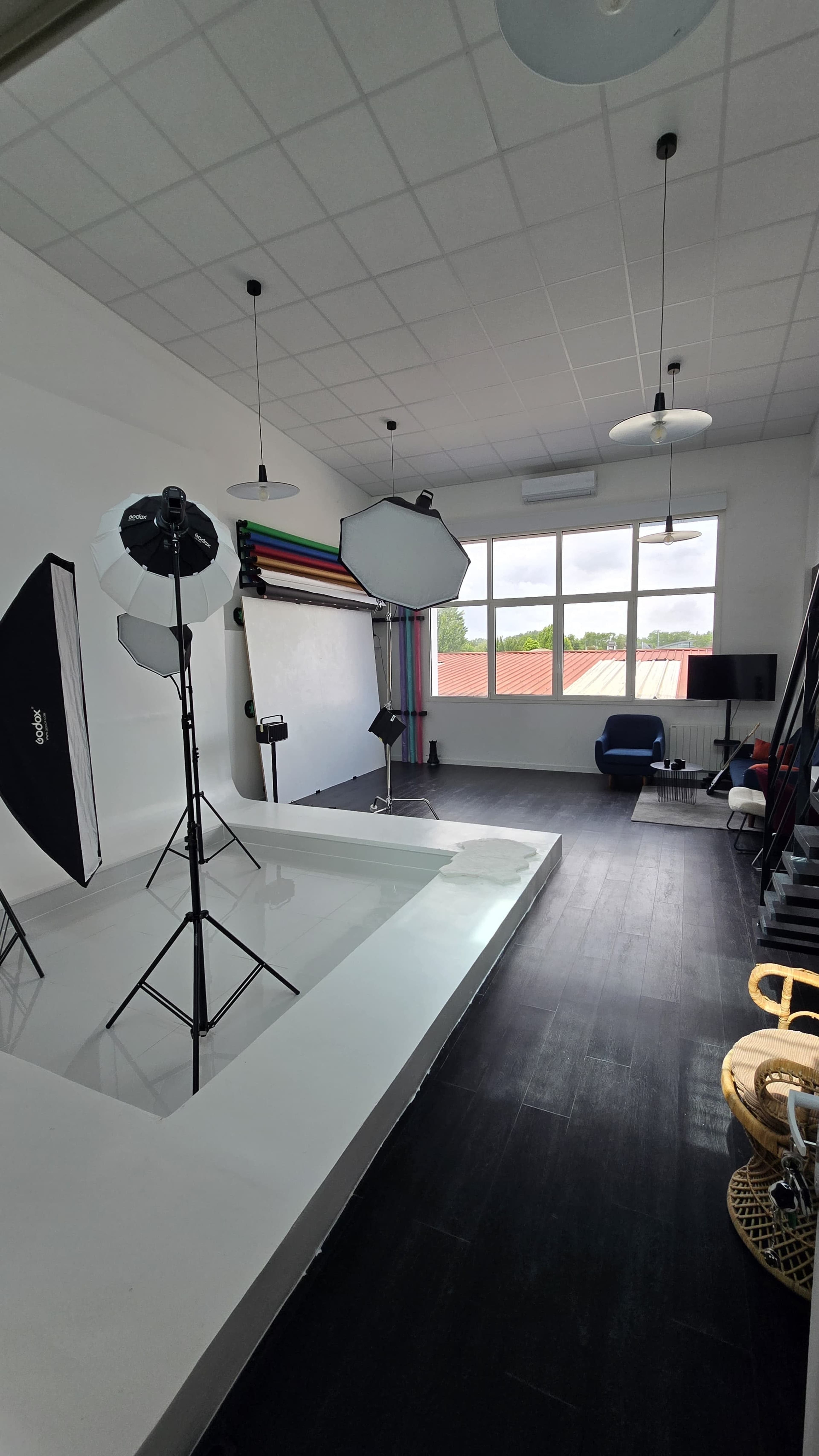 The image shows a photography studio with various lighting equipment, a seamless backdrop, and large windows allowing natural light to enter.