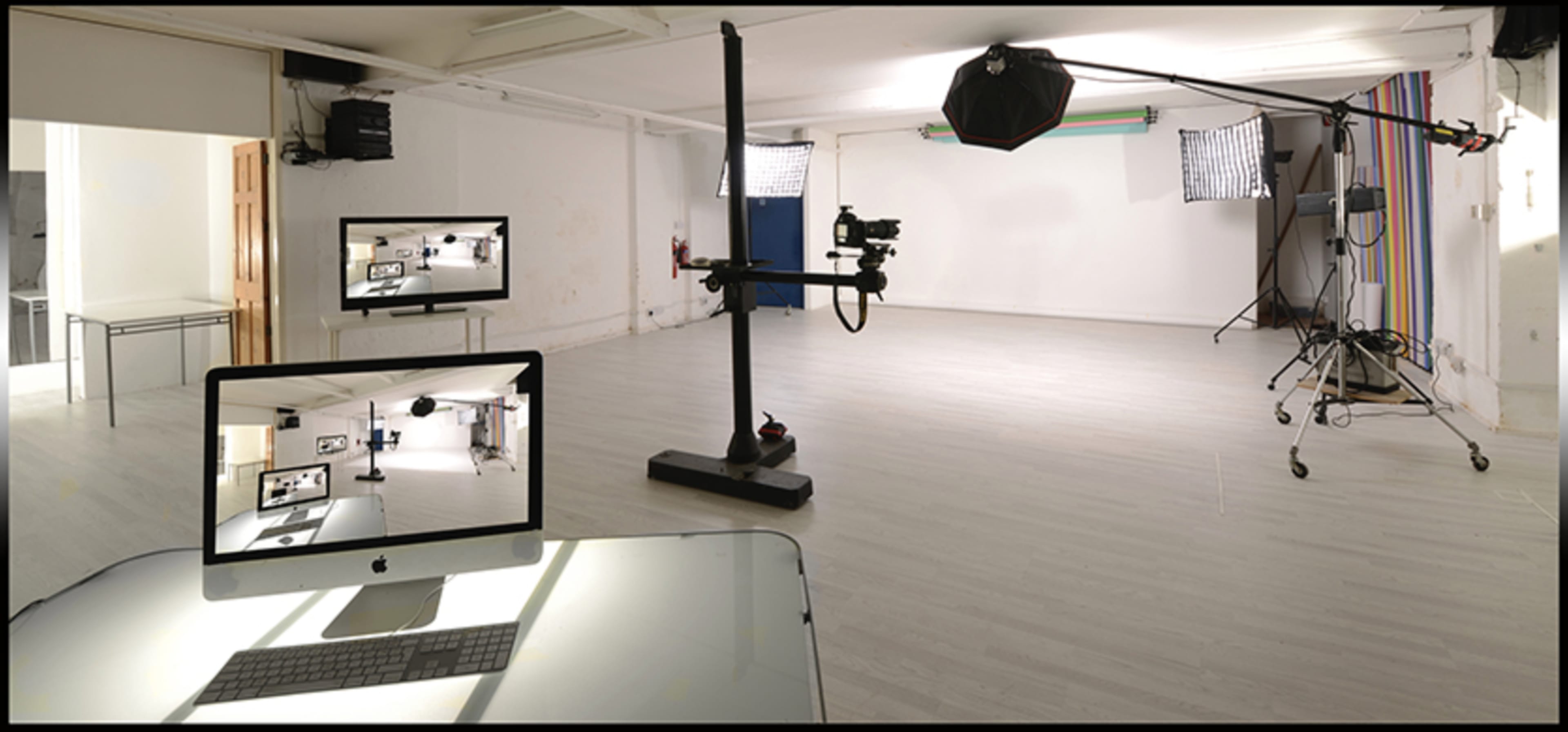 A photography studio is equipped with lighting equipment, a camera on a stand, and a computer displaying a live feed of the space.