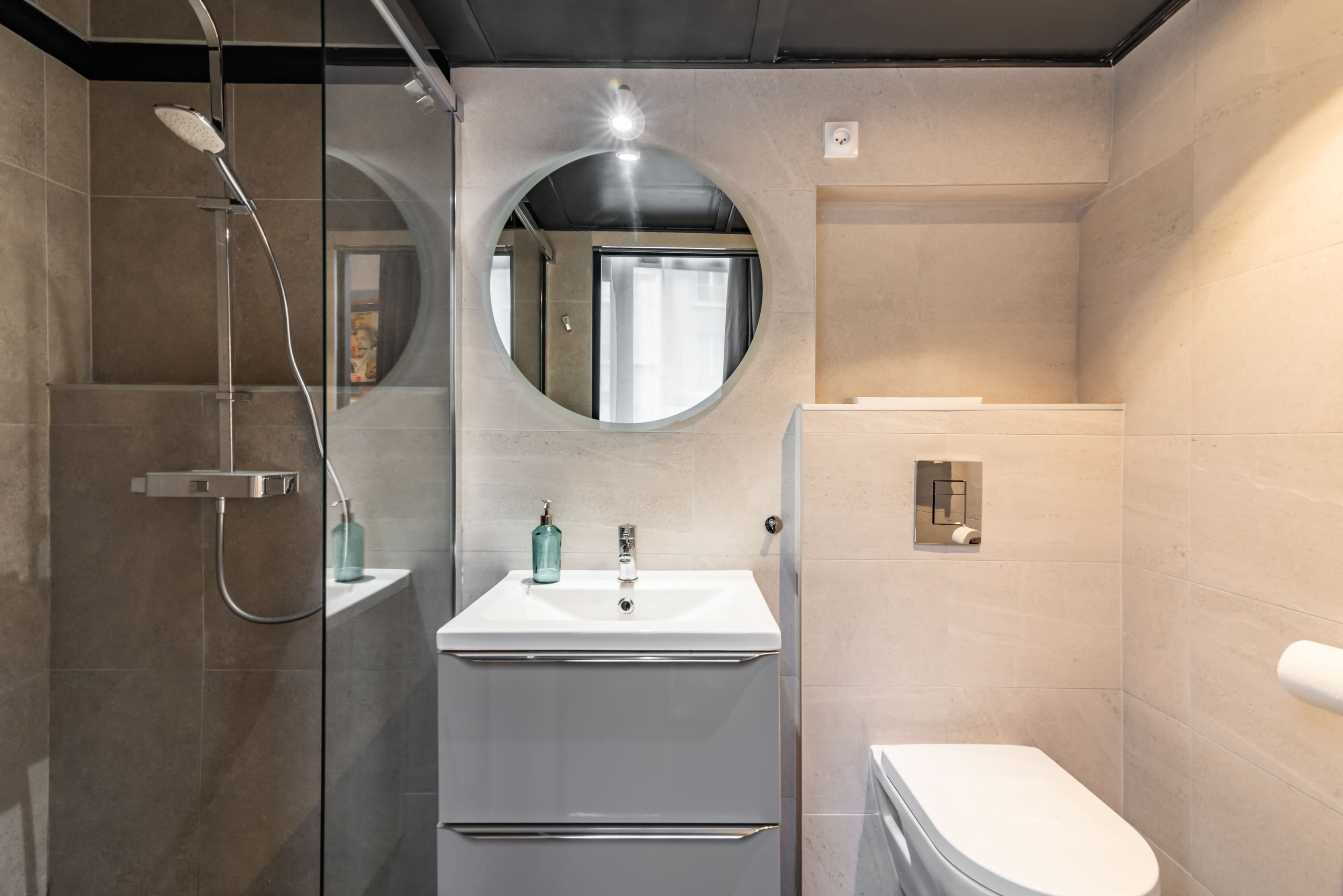 A modern bathroom featuring a shower with glass walls, a round mirror above a white sink, and a toilet beside a minimalist cabinet.
