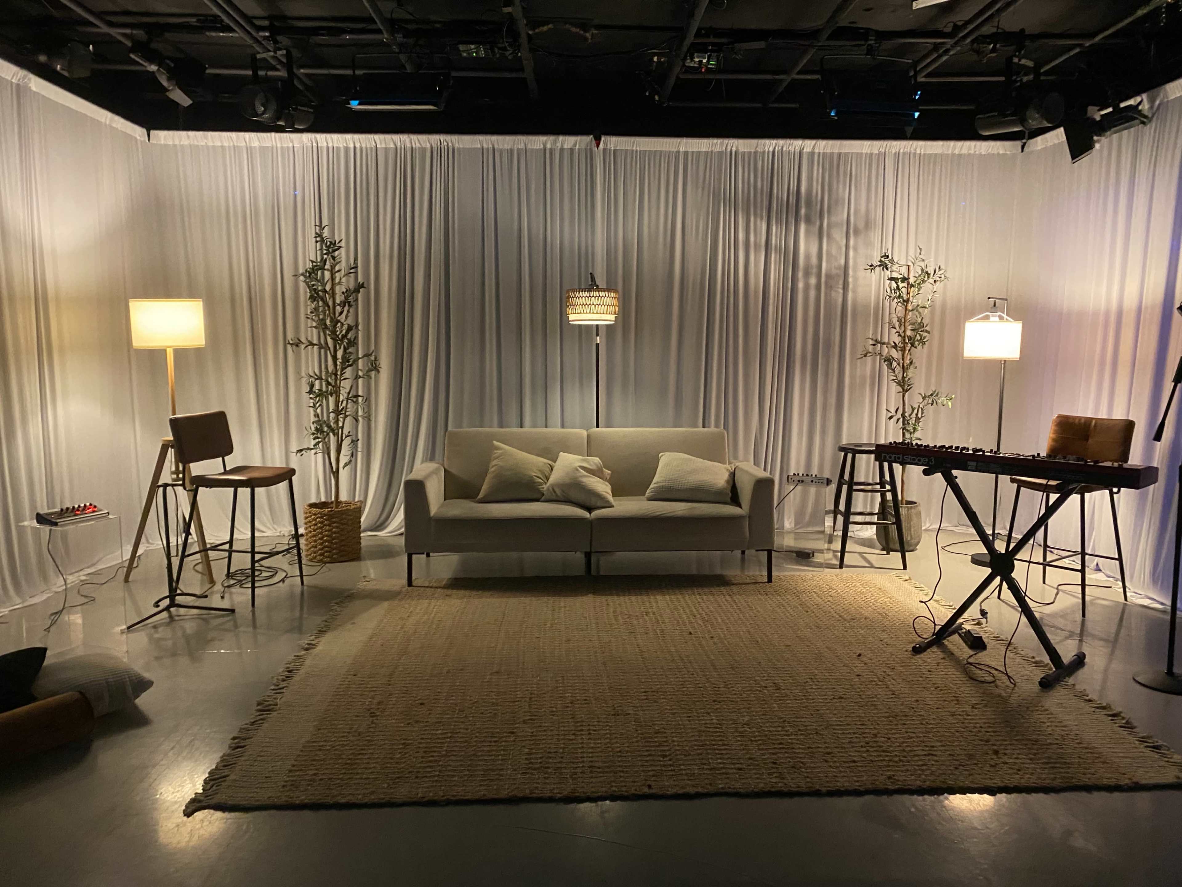 The scene features a minimalist studio setup with a couch, two lamps, a small table, a keyboard, and decorative plants against a backdrop of white drapery.